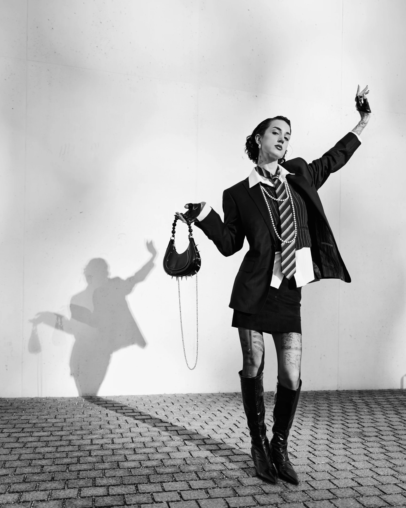In this striking black-and-white portrait, a confident woman stands against a stark, minimalist concrete wall, her silhouette sharply defined by dramatic lighting. Her dark, styled hair frames a poised face with a subtle, defiant expression, one hand raised high in a gesture of power or rebellion while the other holds a spiked, crescent-shaped handbag with a chain strap. She wears a tailored black blazer over a crisp white collared shirt, layered with a striped tie and multiple strands of pearls, creating a juxtaposition of formal and edgy aesthetics. Her outfit is completed with a short skirt, sheer patterned tights revealing tattoos on her legs, and glossy knee-high boots, reinforcing a...