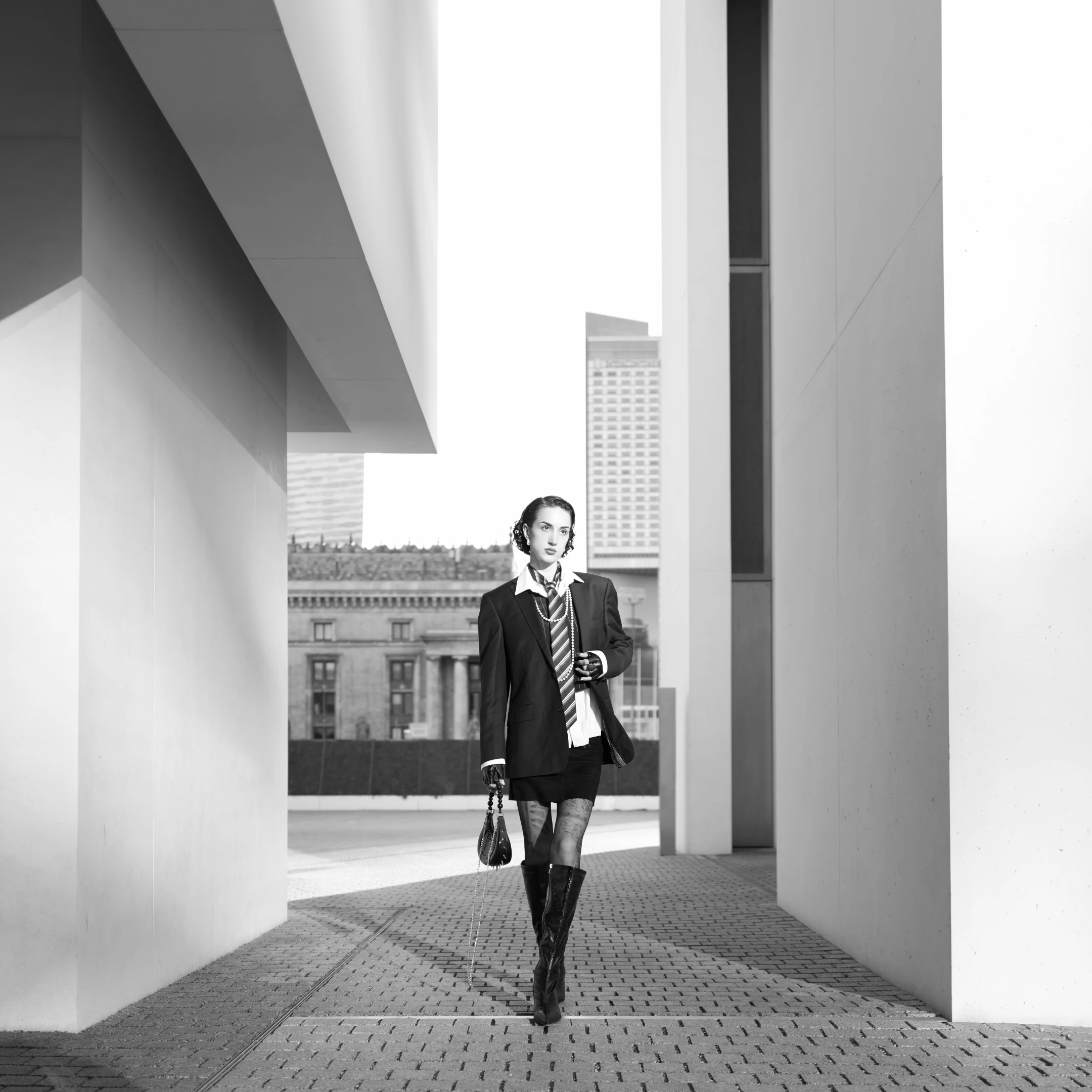 In this striking black-and-white portrait, a woman strides confidently through a modern architectural corridor, framed by stark white concrete pillars and a high-contrast sky. Her expression is poised and direct, gazing forward with a calm, almost defiant intensity, suggesting a sense of purpose or authority. She wears a tailored black blazer over a crisp white shirt, accessorized with a striped tie and layered pearl necklaces, exuding a vintage yet contemporary elegance. Her ensemble is completed with a short skirt, sheer tights, and tall black boots, adding a touch of boldness to her otherwise refined look. She carries a small, structured handbag with a chain strap, held loosely in one ...