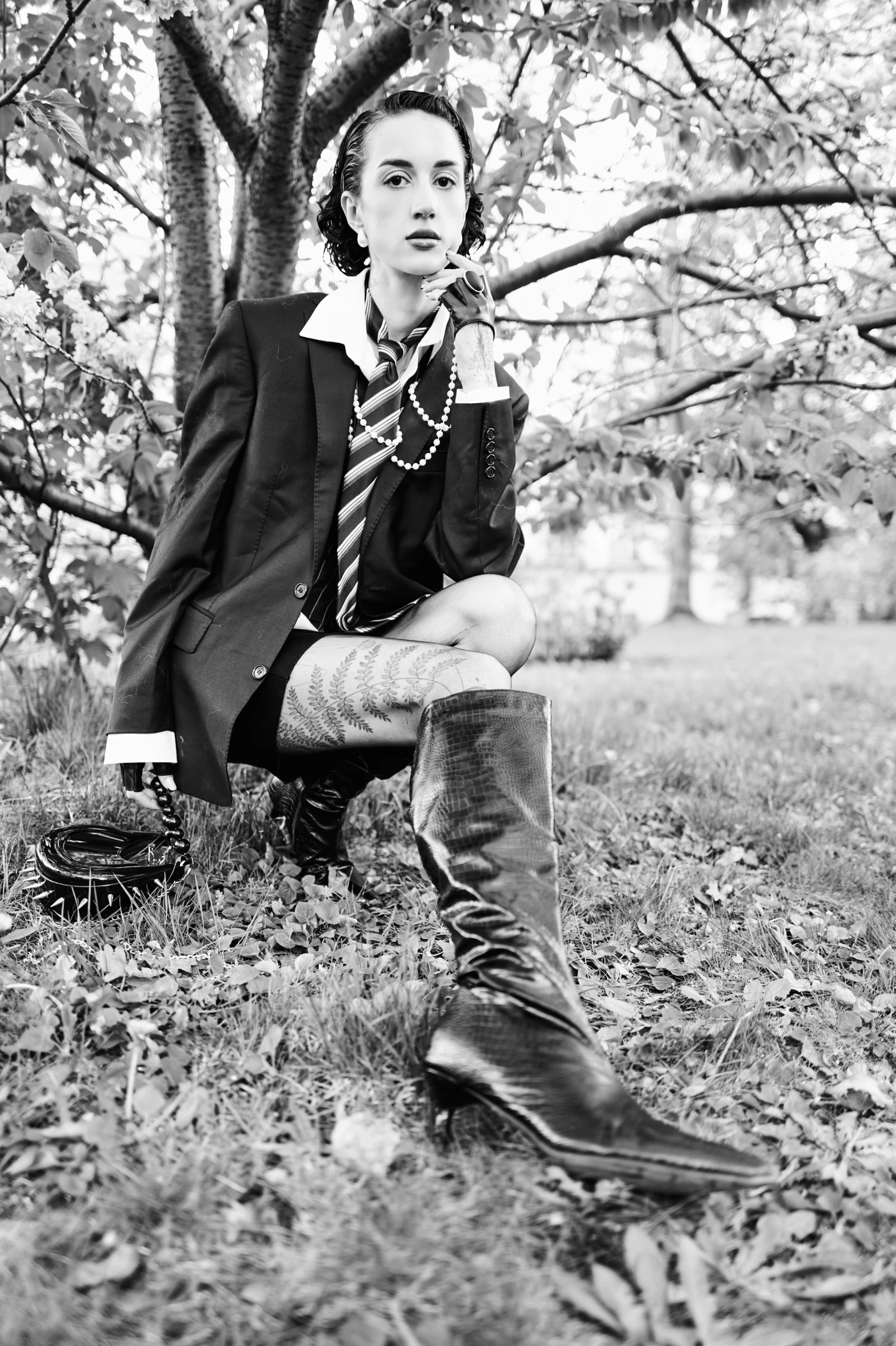 In this striking black-and-white portrait, a young woman with dark, wavy hair styled in a vintage fashion sits confidently on the grass beneath a blossoming tree, her gaze fixed directly at the viewer with an air of quiet intensity. Her expression is composed yet enigmatic, with one hand delicately resting near her chin, suggesting contemplation or defiance. She wears a tailored blazer over a crisp white-collared shirt, paired with a striped tie and layered pearl necklaces, evoking a formal, almost academic aesthetic that contrasts sharply with her bold, high-heeled crocodile-patterned boots. Her left thigh displays a detailed fern tattoo, adding an element of nature and personal artistry...