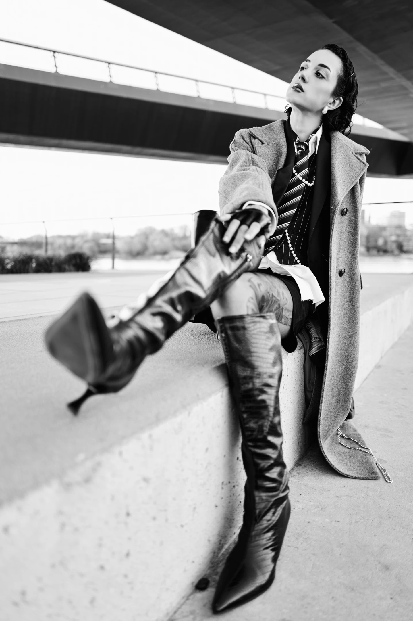 In this striking black-and-white portrait, a woman exudes confidence and drama, seated on a concrete ledge under an architectural overpass. Her dark hair is pulled back elegantly, accentuating her bold expression as she gazes upward with a poised, almost defiant look. She wears a long, textured overcoat draped over a layered ensemble featuring a striped tie, a crisp white shirt, and a dark vest, suggesting a blend of formal and edgy fashion. Her legs are dramatically crossed, one knee lifted high, revealing a tattooed thigh and a pair of glossy, high-heeled thigh-high boots that gleam under the stark lighting. The lighting is high-contrast, casting sharp shadows and emphasizing the textur...