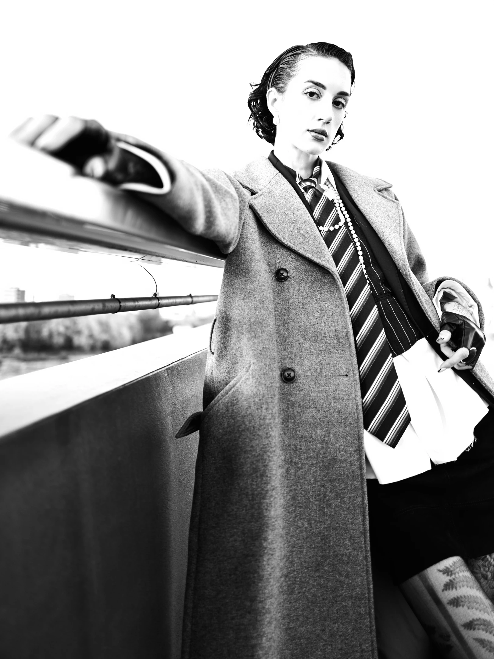 This is a striking black-and-white portrait of a woman standing confidently on a bridge railing, her gaze fixed directly at the viewer with an air of quiet intensity. Her dark, wavy hair is neatly styled, framing a face adorned with subtle makeup, including defined eyes and a bold lip. She wears a long, textured overcoat with a double-breasted design, layered over a formal vest and a striped tie, suggesting a vintage or retro-inspired fashion aesthetic. Her left hand rests on the railing, while her right hand holds a white garment or cloth, adding a sense of narrative or mystery to the image. The lighting is high-contrast, with bright exposure on the subject’s face and upper body, creatin...