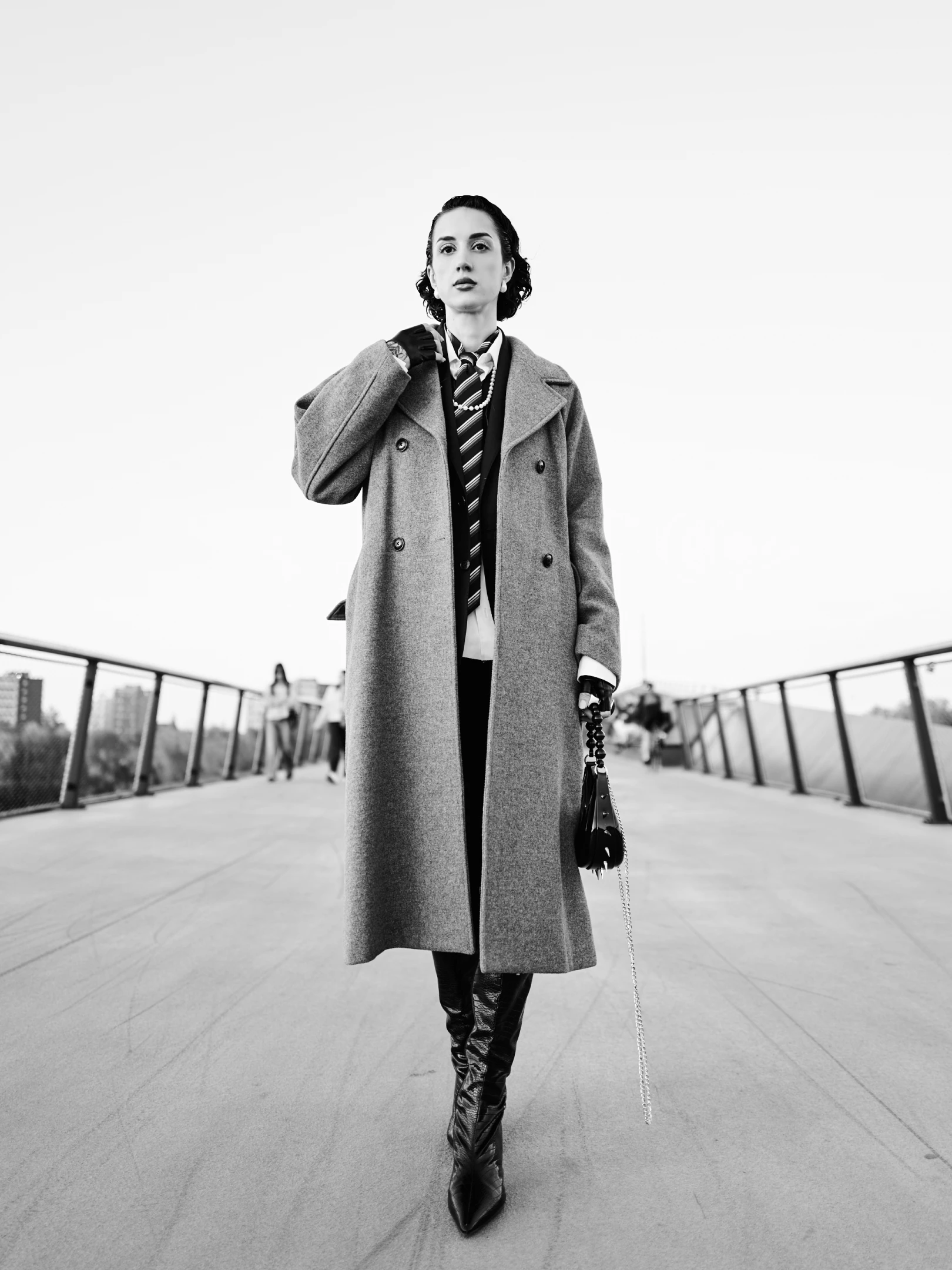 The portrait captures a poised, confident woman standing center-frame on a wide, open pedestrian bridge under a bright, overcast sky. Her dark, wavy hair is neatly styled, framing a face with a direct, unflinching gaze that conveys strength and quiet authority. She wears a long, textured gray overcoat, layered over a crisp white shirt, a striped tie, and a dark vest, suggesting a deliberate, fashion-forward blend of classic and edgy styles. Her left hand rests on her coat’s shoulder, while her right hand holds a small, studded black handbag with a chain strap, adding a touch of modern luxury. She wears tall, glossy black boots that accentuate her commanding presence. The lighting is soft ...