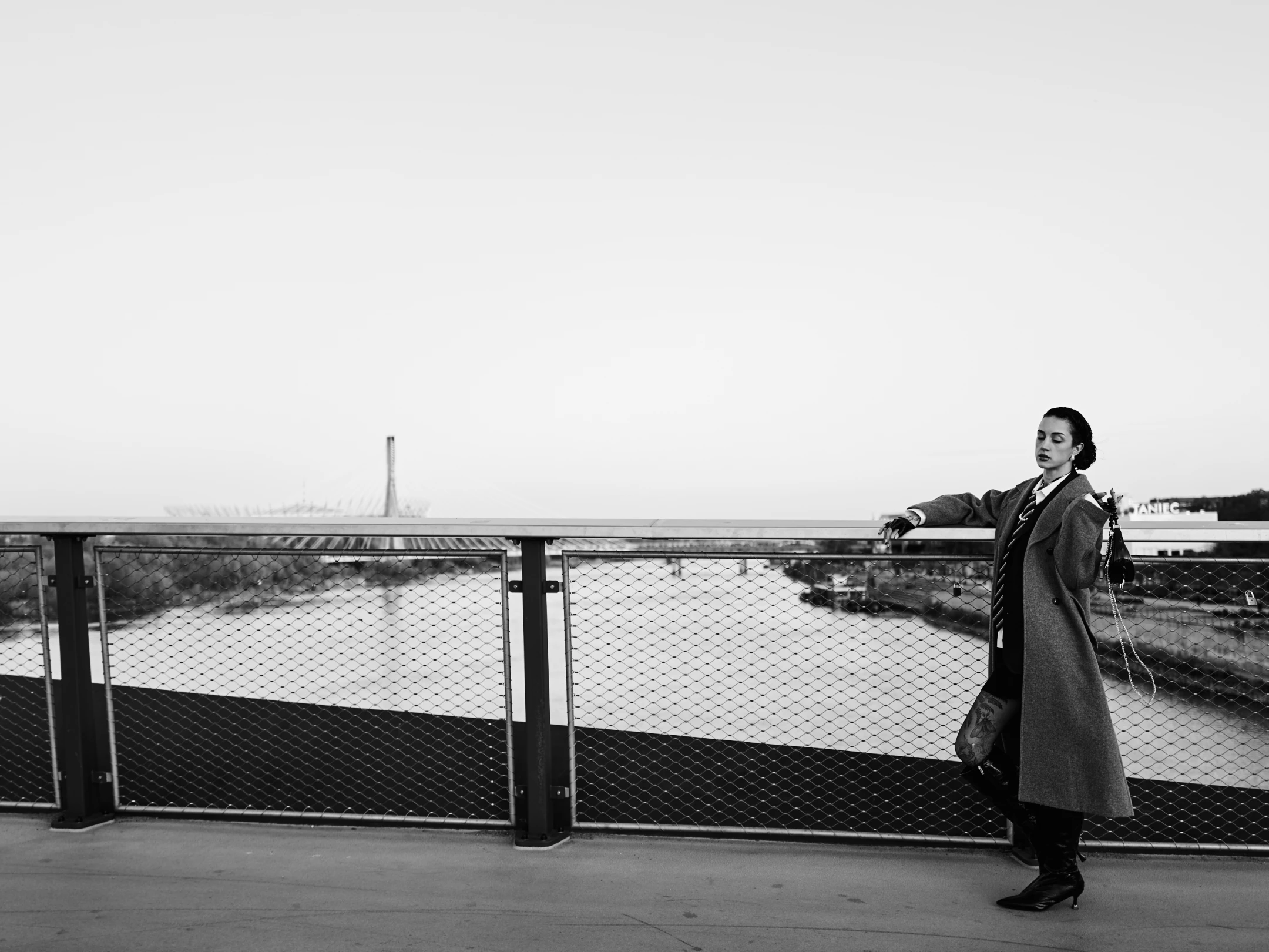 In this striking black-and-white portrait, a woman stands confidently on a modern bridge, leaning against a chain-link fence that separates her from a wide river below. Her dark hair is neatly pulled back, accentuating her poised expression as she gazes thoughtfully toward the horizon, her lips slightly parted and eyes softly focused. She wears a long, textured overcoat that drapes elegantly over a dark outfit, complemented by high-heeled boots and a small, chain-handled bag slung over her shoulder. Her left arm rests casually on the railing, while her right hand, adorned with a tattooed forearm, hangs loosely by her side, suggesting a relaxed yet deliberate stance. The lighting is soft a...
