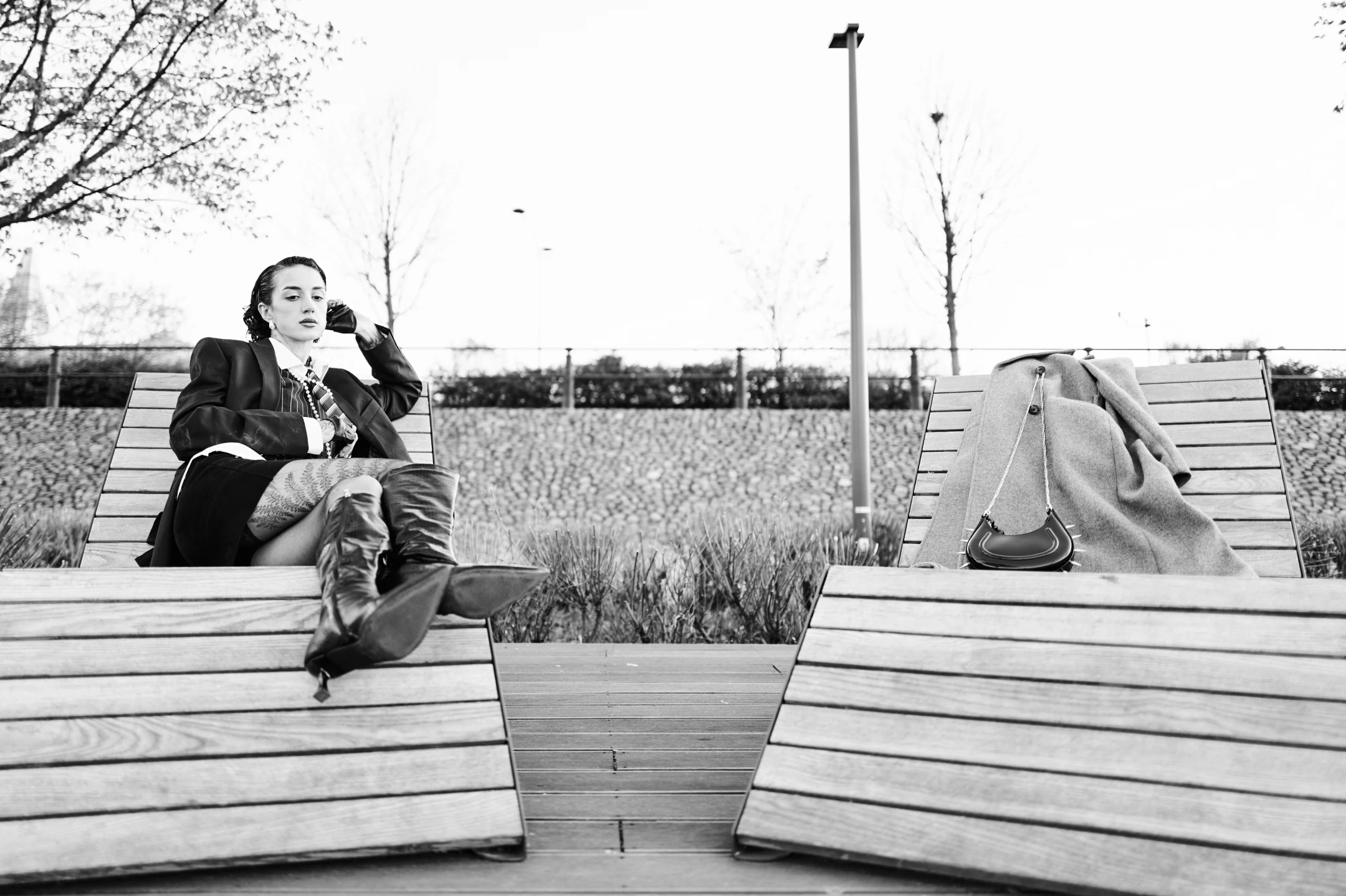 In this striking black-and-white portrait, a woman sits poised on a modern wooden bench, exuding confidence and quiet contemplation. Her dark hair is neatly pulled back, accentuating her strong jawline and piercing gaze as she looks off into the distance, one hand resting thoughtfully near her ear. She wears a structured blazer over a striped shirt and tie, paired with a short skirt and tall, glossy boots that add an edgy, fashion-forward flair. Her expression is calm yet introspective, suggesting a moment of pause or reflection. The lighting is soft and even, likely from an overcast sky, lending a timeless, almost cinematic quality to the image. Behind her, a minimalist park setting unfo...