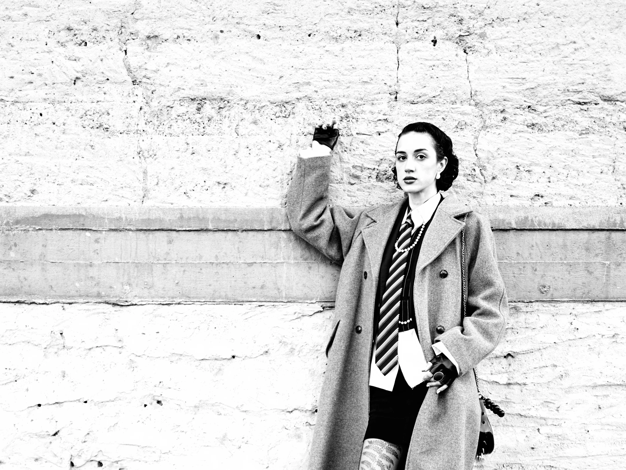 In this striking black-and-white portrait, a woman stands confidently against a textured, weathered concrete wall, her dark hair styled in a polished updo that frames her face. Her expression is poised and direct, with a neutral gaze fixed toward the camera, suggesting a sense of calm authority. She wears a long, double-breasted overcoat, layered over a crisp white shirt, a striped tie, and a pearl necklace, creating a juxtaposition of formal elegance and edgy modernity. Her gloved hands—one raised slightly as if gesturing or touching the wall, the other holding a small, dark object—add a touch of deliberate movement to the stillness. The lighting is even and high-contrast, emphasizing th...