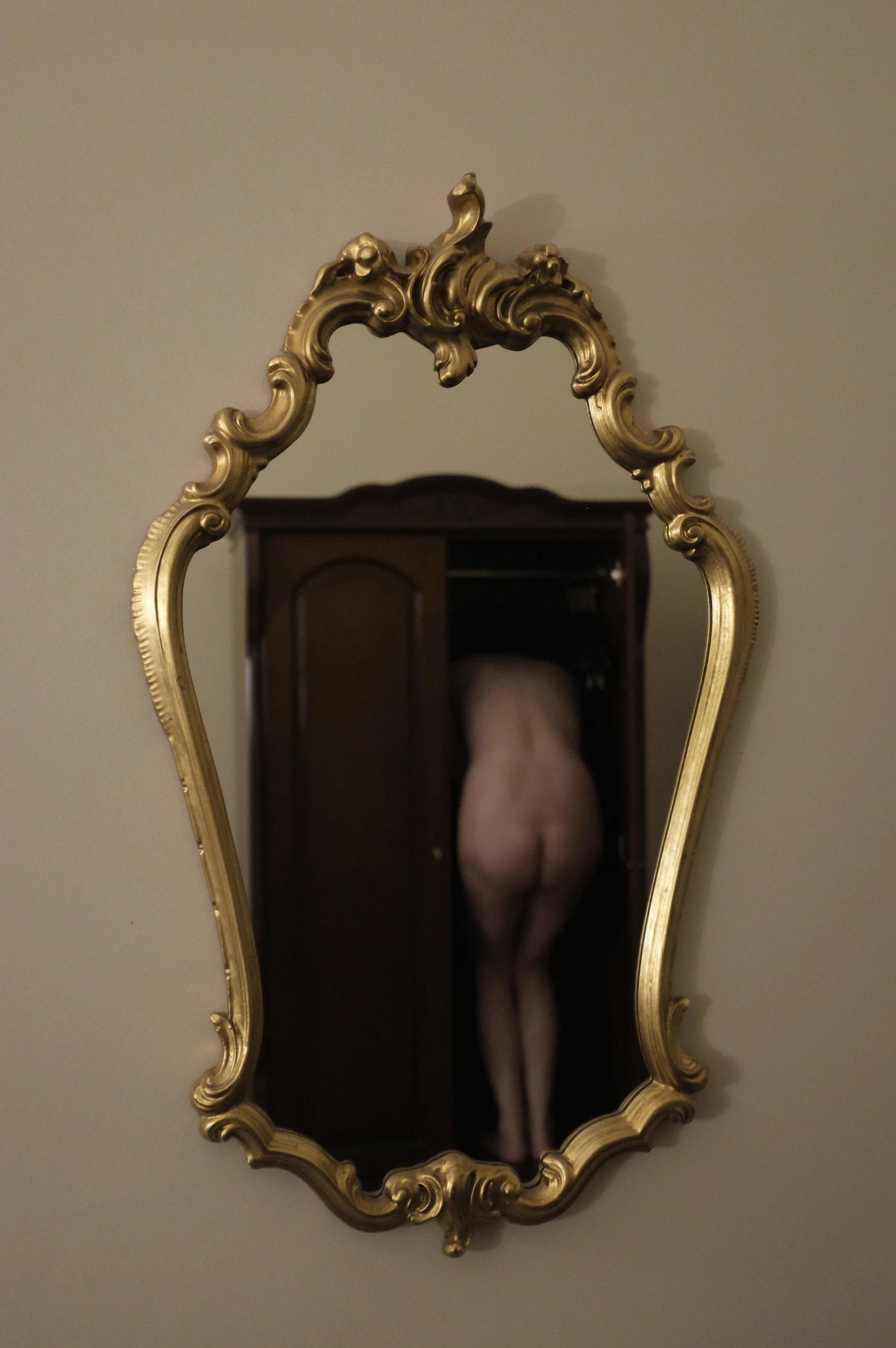 A nude figure stands behind a dark wooden door, partially visible through a large, ornate gold-framed mirror mounted on a beige wall. The mirror's elaborate, baroque-style frame features swirling gold embellishments and a pointed top, reflecting the figure's back and legs as they stand facing away from the viewer. The lighting is dim and warm, casting soft shadows and emphasizing the smooth contours of the body. The mood is intimate and contemplative, with the mirror creating a layered, reflective composition. The background is a plain, neutral-toned wall, keeping focus on the mirror's reflection and the figure's poised, silent stance.