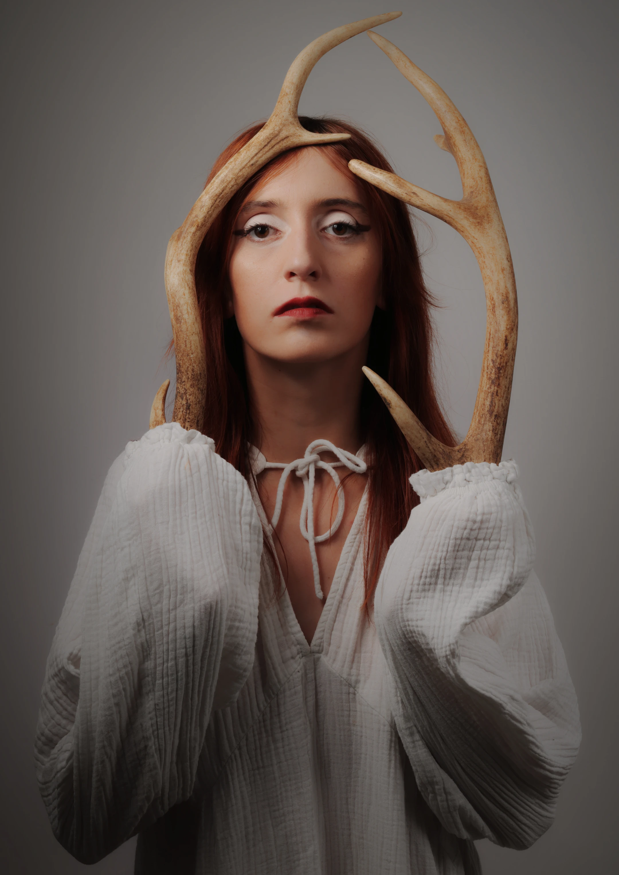A woman with long, reddish-brown hair stands against a neutral gray backdrop, her expression solemn and contemplative as she gazes slightly upward. Large, branching antlers emerge from her head and shoulders, framing her face and extending outward, suggesting a mythical or symbolic presence. She wears a flowing, textured white blouse with a tie-front neckline, adding a soft, ethereal quality to the image. Soft, directional lighting highlights the antlers' natural grain and the woman's facial features, casting gentle shadows that deepen the portrait's introspective mood. The minimalist background emphasizes the subject and her surreal, nature-inspired adornment, evoking themes of wildernes...