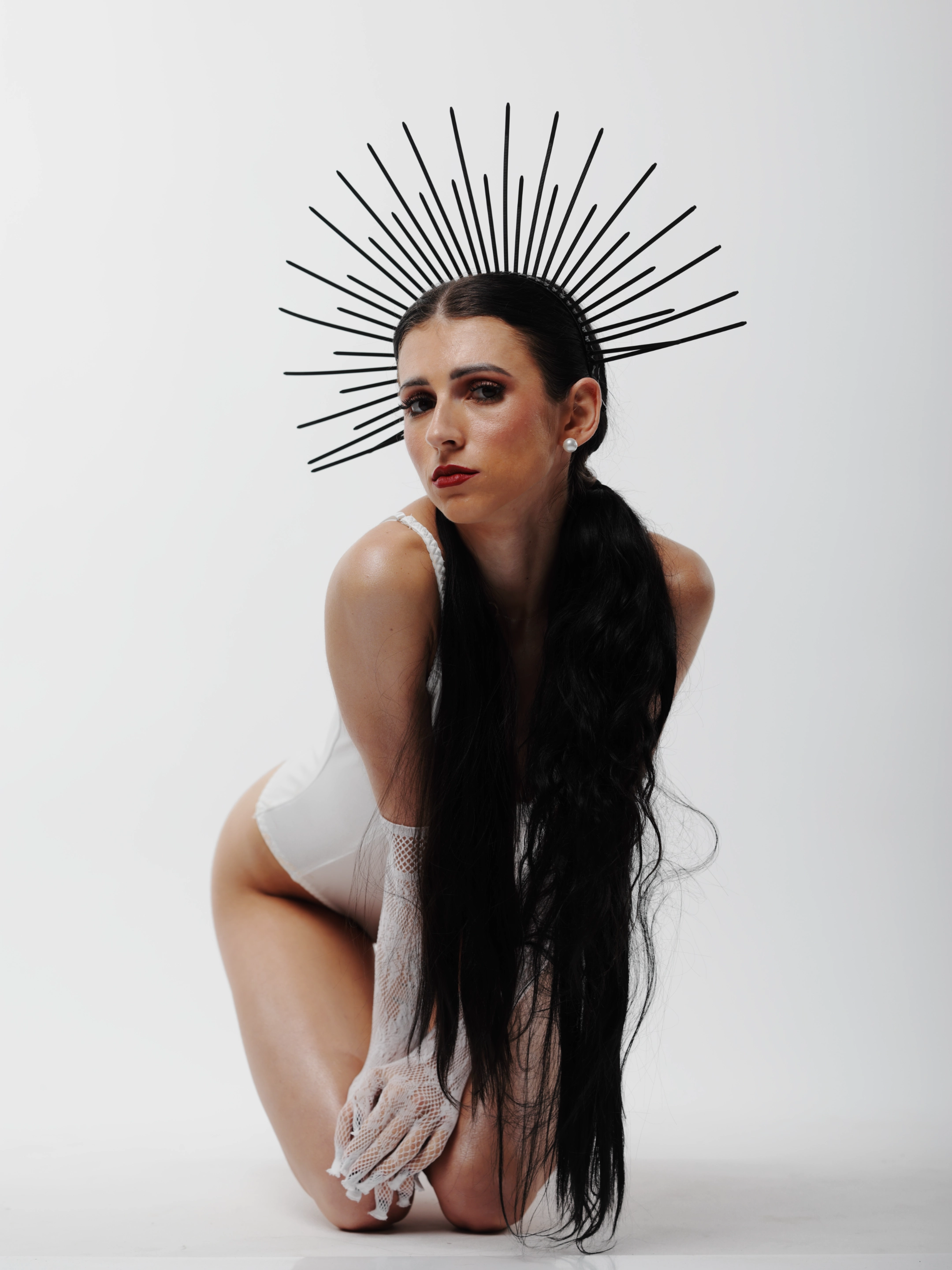 A woman with long, dark, wavy hair kneels on a white surface, wearing a white bodysuit and lace gloves, with a striking black spiked crown radiating behind her head. Her expression is intense and direct, gazing forward with bold red lips and dark eye makeup. Soft studio lighting highlights her features and the texture of her attire, while the stark white background emphasizes her dramatic, almost mythic presence. The mood is surreal and powerful, blending elegance with an edgy, otherworldly aesthetic.