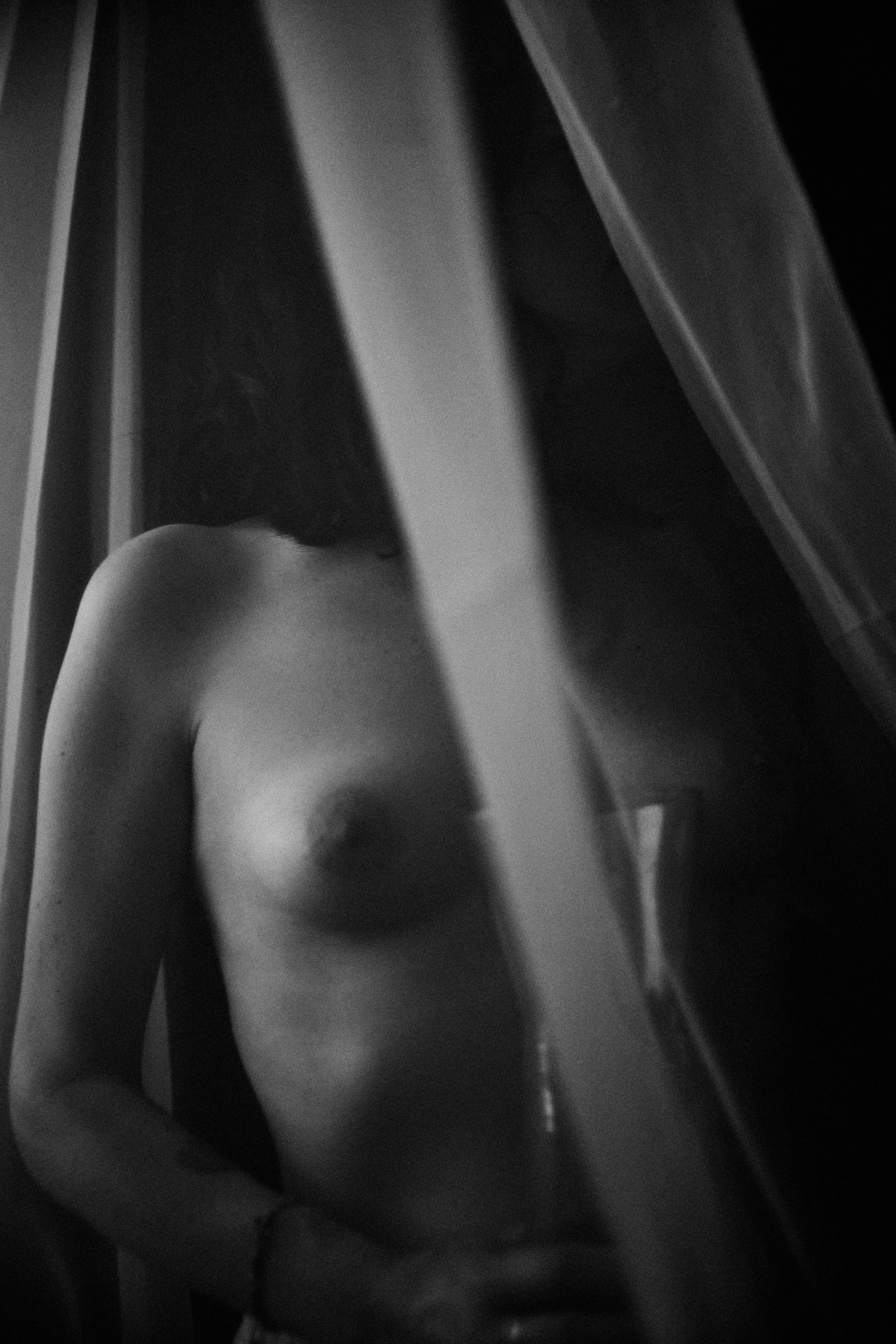 A black-and-white portrait captures a nude figure partially obscured by heavy, draped fabric, likely curtains, creating a sense of intimacy and mystery. The subject's upper body is visible, with arms gently crossed over the torso, suggesting a quiet, contemplative pose. Soft, directional lighting highlights the curves of the body while leaving much of the scene in shadow, evoking a moody, sensual atmosphere. The background is dark and undefined, emphasizing the figure and the fabric's texture, which adds depth and a tactile quality to the image. The overall mood is private, evocative, and emotionally charged, focusing on form and shadow rather than explicit detail.