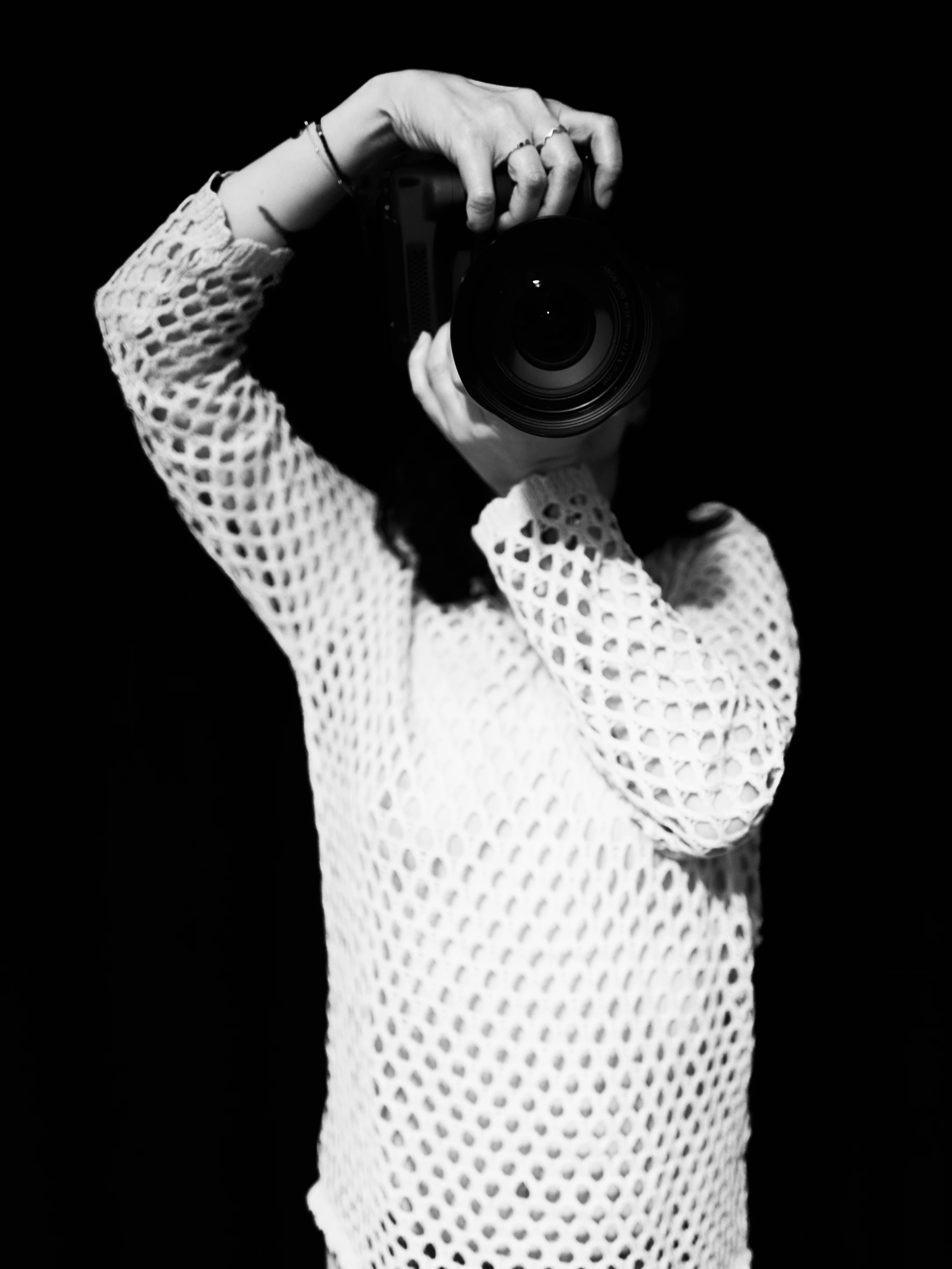 A woman in a white, open-knit, mesh-like sweater stands against a stark black background, holding a professional camera with both hands, obscuring her face. Her pose is focused and deliberate, suggesting she's capturing a shot or embodying the act of photography. The lighting highlights the texture of her sweater and the camera's details, creating a high-contrast, minimalist mood. The black background isolates her, emphasizing the interplay of light and shadow, evoking a sense of mystery and artistic intent.