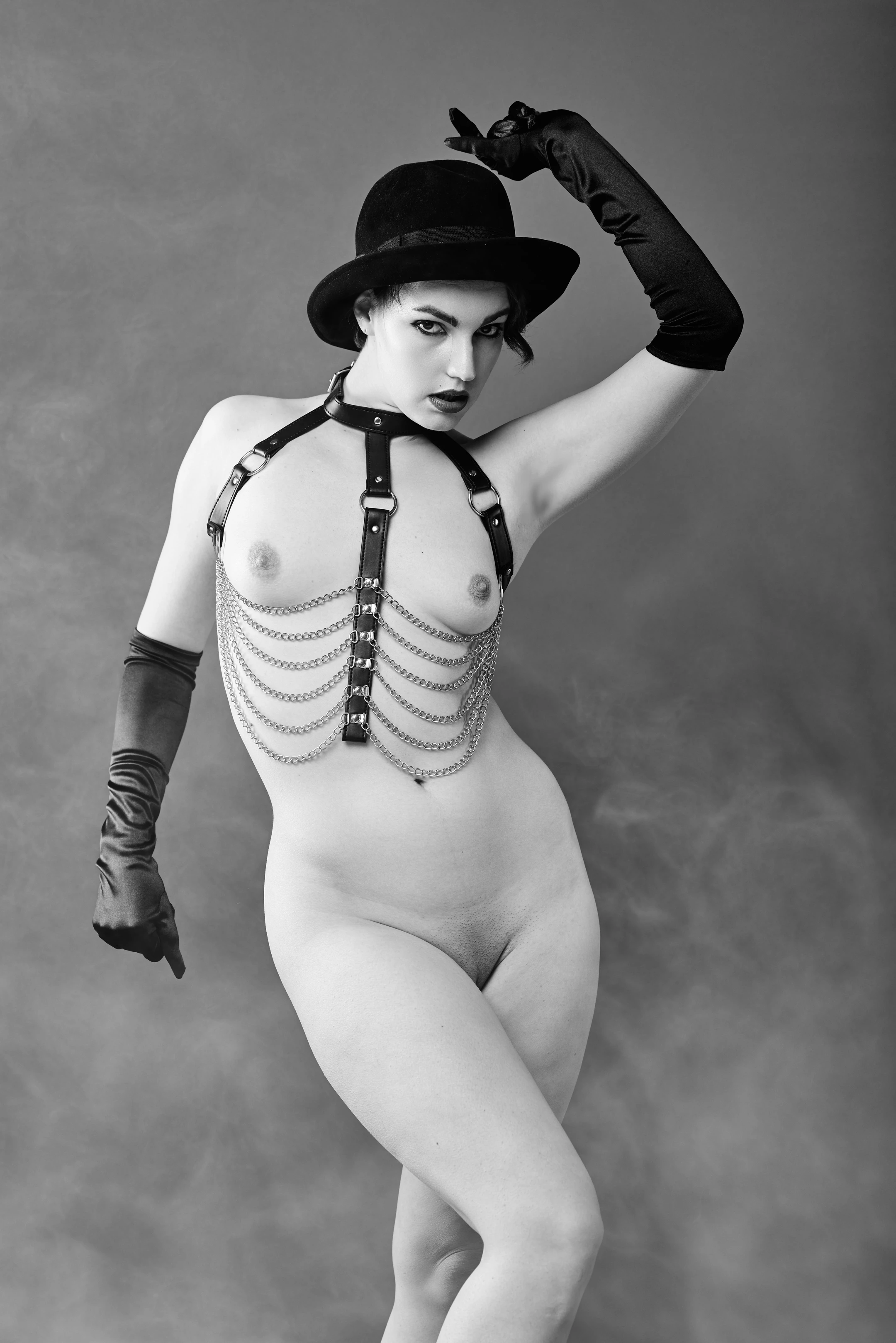 A black-and-white portrait of a woman posing confidently with a direct gaze, wearing a black velvet fedora, long black gloves, and a leather harness adorned with cascading silver chains. Her body is nude except for the harness and gloves, emphasizing her form against a soft, neutral gray backdrop. Lighting highlights her silhouette and the textures of the leather and chains, creating a moody, stylized aesthetic. The pose is dynamic, with one arm raised and the other pointing downward, conveying power and elegance.