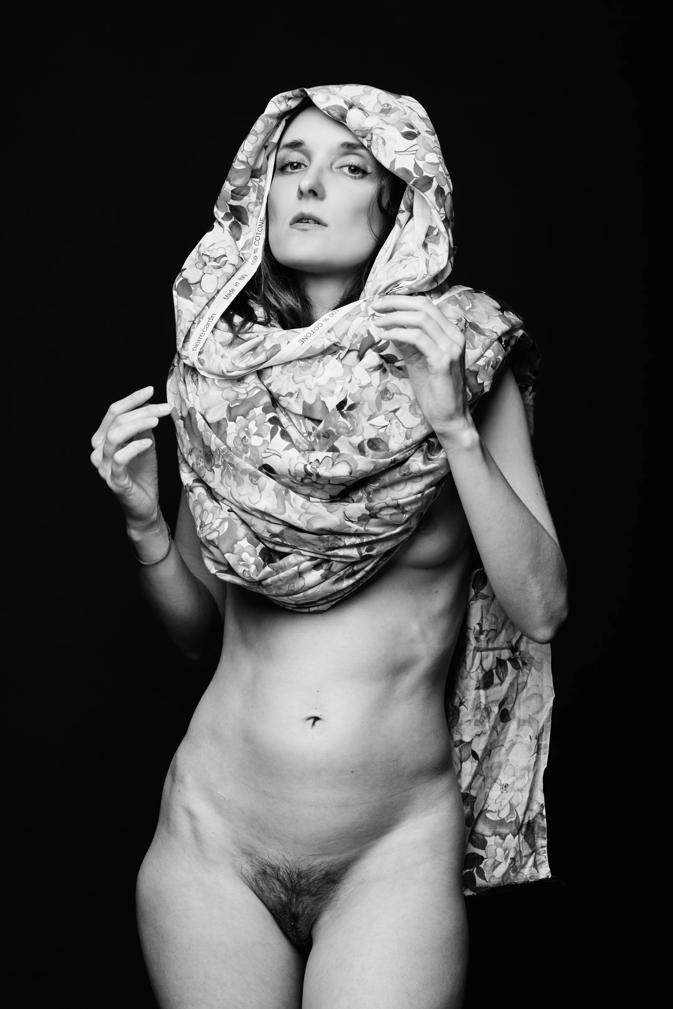 A striking black-and-white portrait of a nude woman standing against a stark, deep black background, which isolates her form and emphasizes the texture and contours of her body. Her expression is calm and contemplative, with eyes gazing slightly upward and her lips slightly parted, suggesting a quiet introspection or vulnerability. She is draped in a large, floral-patterned scarf, branded Pierre Cardin and Made in Italy, which covers her head and cascades down her torso, partially concealing her body while leaving her abdomen, hips, and legs exposed. The scarf's delicate floral design contrasts with the starkness of the background and the rawness of her nudity, creating a juxtaposition of elegance and vulnerability.