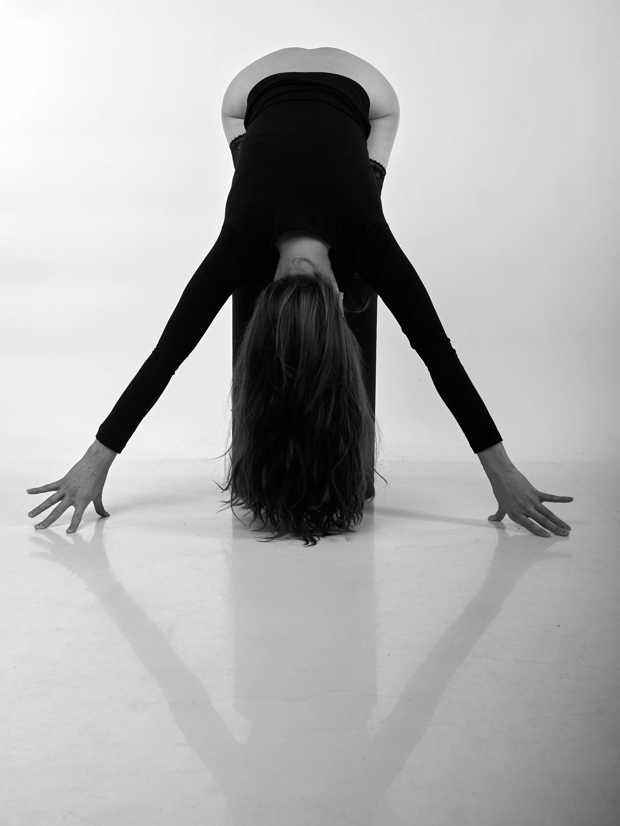 A striking black-and-white portrait of a woman captured in a dynamic, inverted yoga pose, likely a downward dog variation, with her body arched backward and arms extended toward the floor. Her long, dark hair cascades down toward the reflective white floor, creating a dramatic contrast against the stark, minimalist background. She wears a form-fitting black long-sleeve top and matching leggings, with delicate lace trim at the waistline, adding a touch of elegance to the athletic posture. Her expression is neutral, focused, and serene, suggesting deep concentration or meditative calm. The lighting is soft and even, illuminating her silhouette with clarity while casting subtle shadows on the smooth surface beneath her.