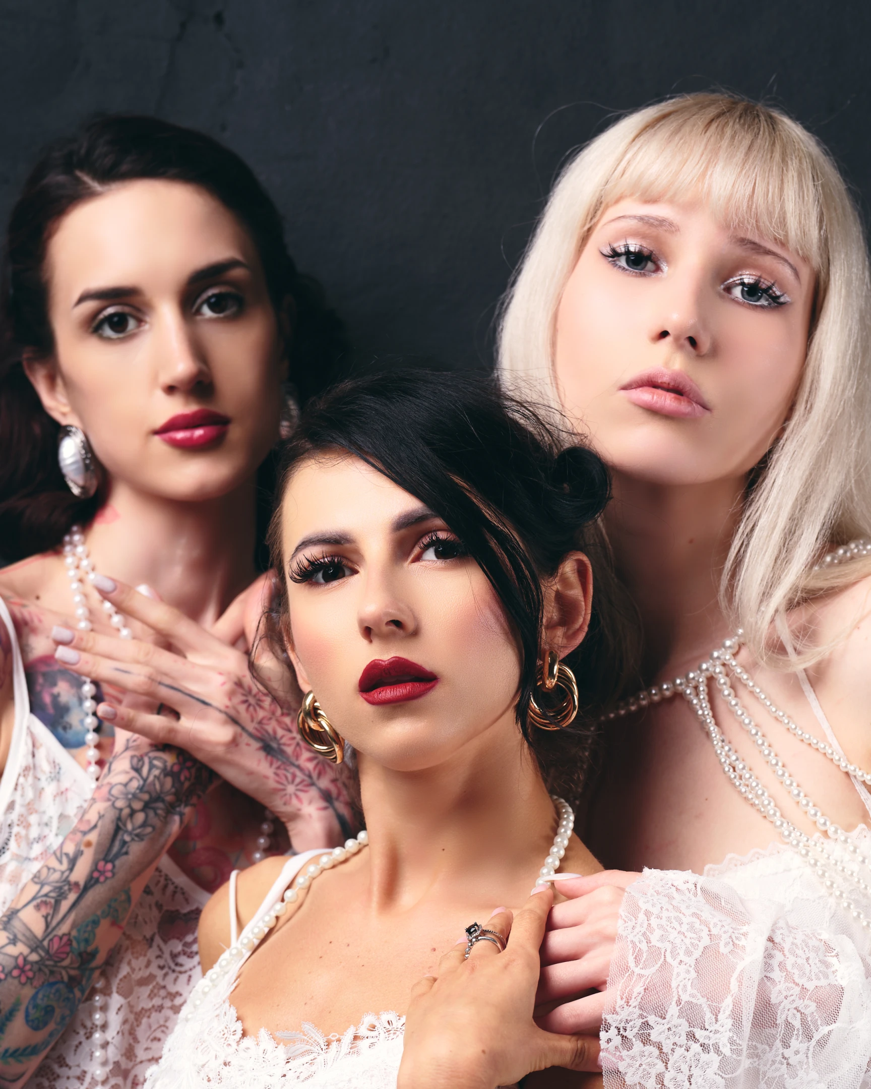 This portrait features three women posing closely together against a dark, textured backdrop that enhances the vibrancy of their attire and makeup. The central figure, with dark hair and bold red lipstick, gazes directly at the viewer with a confident, neutral expression, adorned in a delicate white lace top and a pearl necklace, her hand gently resting on her chest. To her left, a woman with dark hair and tattoos on her arms wears a white lace garment and large silver earrings, her hand placed over the central figure’s shoulder, displaying a calm, composed demeanor. On the right, a blonde woman with straight bangs and dramatic eyeliner wears a white lace dress layered with multiple pearl...
