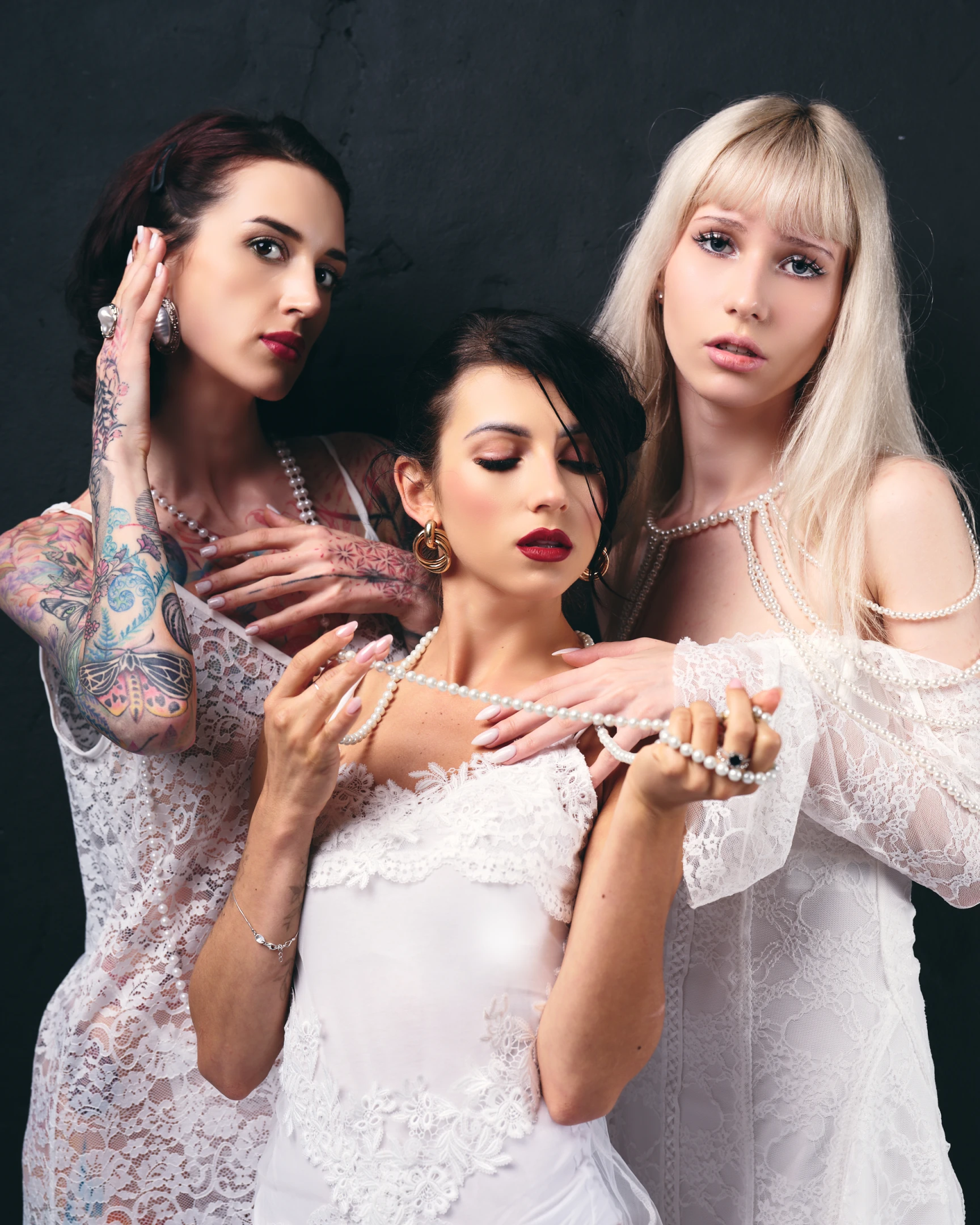 This portrait features three women in a striking, high-contrast composition against a dark, textured backdrop that emphasizes their luminous white lace attire. The central figure, with dark hair and closed eyes, wears a delicate white lace dress adorned with pearls and gold hoop earrings, exuding a serene, almost ethereal calm as she gently holds a pearl necklace. To her left, a woman with deep red hair and visible tattoos on her arms and hands gazes directly at the camera with a bold, confident expression, her fingers near her ear and a pearl necklace resting on her shoulder. On the right, a blonde woman with straight bangs and a soft, neutral expression wears a sheer lace gown embellish...