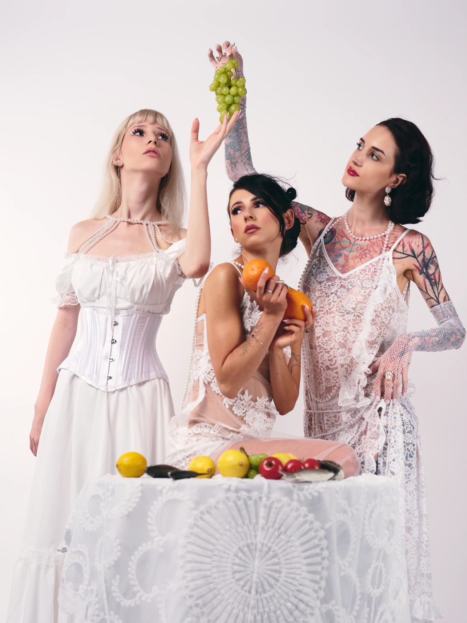 This portrait features three women in an elegant, surreal composition against a stark white background, evoking a vintage yet modern aesthetic. The woman on the left, with platinum blonde hair and bold makeup, wears a structured white corset dress adorned with pearl chains, her gaze upward as if in reverie or anticipation. The central figure, with dark hair and visible tattoos, holds two oranges and wears a sheer lace gown, her expression contemplative as she looks toward the fruit. The woman on the right, with dark wavy hair and red lipstick, sports a lace overlay dress with mesh sleeves and visible tattoos, her hand raised to hold a cluster of green grapes, gazing upward with a poised, ...