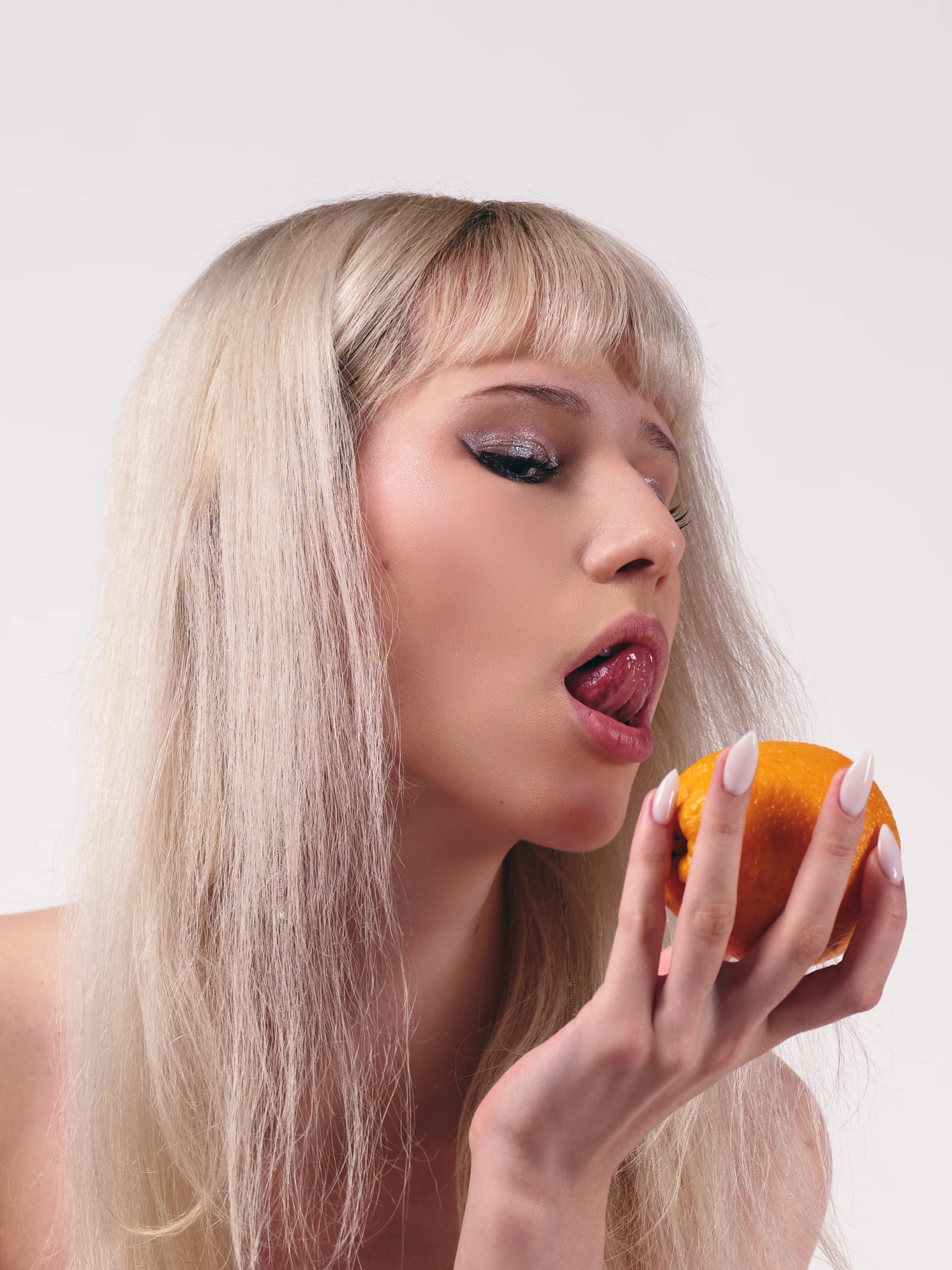 The portrait captures a young woman with long, straight platinum blonde hair and a blunt, even fringe that frames her face. Her makeup is bold and glamorous, featuring shimmering silver eyeshadow, dramatic black winged eyeliner, and glossy pink lips, with her tongue slightly protruding as if tasting or about to bite into a bright orange she holds delicately in her right hand. Her nails are long, manicured, and painted a soft white, adding a clean, polished aesthetic. She is bare-shouldered, suggesting a minimalist or nude top, emphasizing her skin tone and the natural texture of her hair. The lighting is soft and diffused, likely studio-based, casting gentle highlights on her face and the...