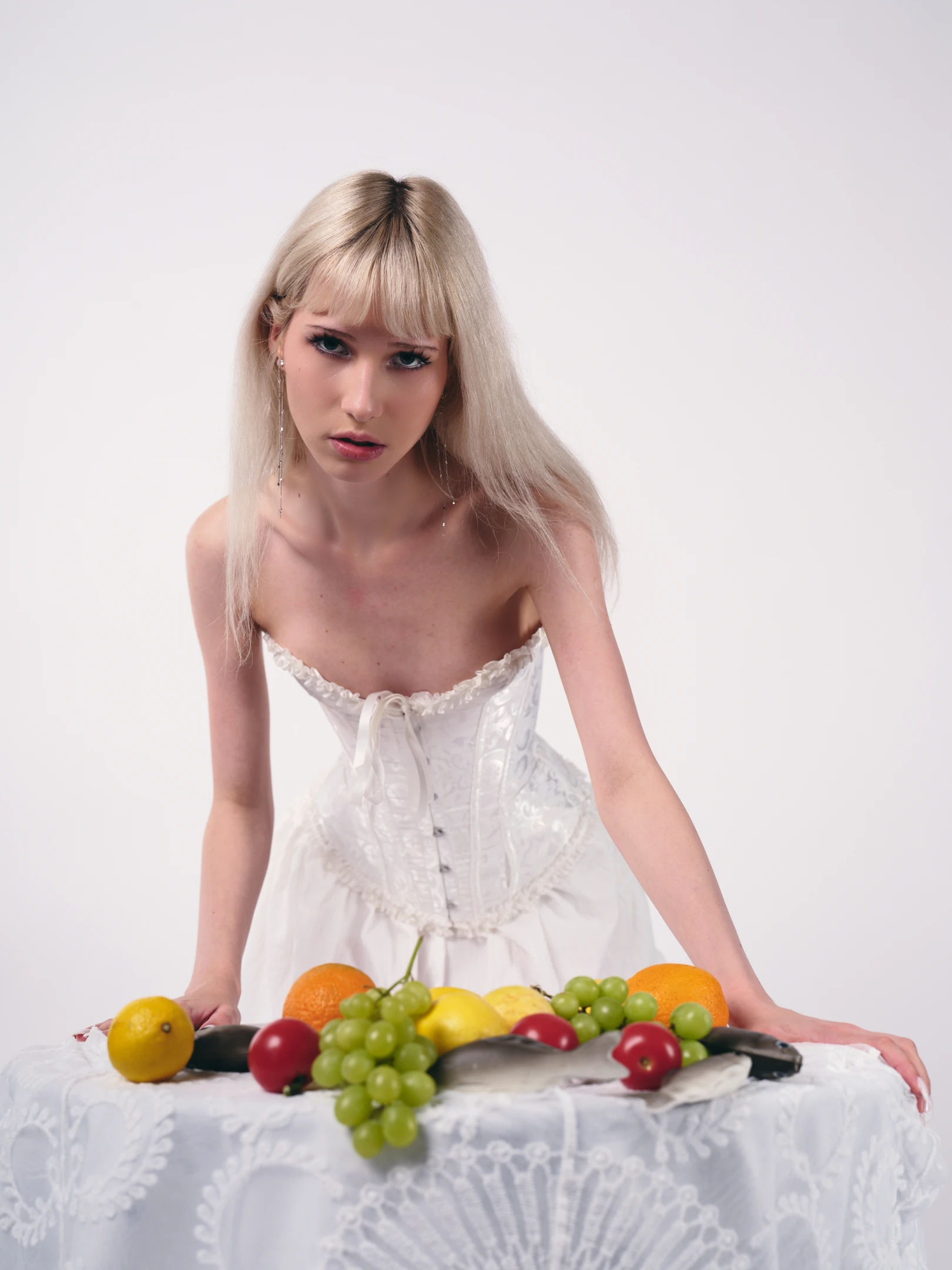 A young woman with long, straight platinum blonde hair and a blunt fringe gazes directly at the camera with a serious, almost somber expression, her lips slightly parted and dark, dramatic eyeliner accentuating her piercing gaze. She wears an ornate white corset-style dress with lace trim and a front tie, suggesting a vintage or fantasy aesthetic, paired with long, dangling silver earrings that catch the light. Her posture is poised and deliberate, leaning forward slightly over a white lace-covered table, hands resting gently on its edge, as if presenting or guarding the still life arrangement before her. The table holds a vibrant assortment of fruit — lemons, oranges, grapes, and tomatoe...