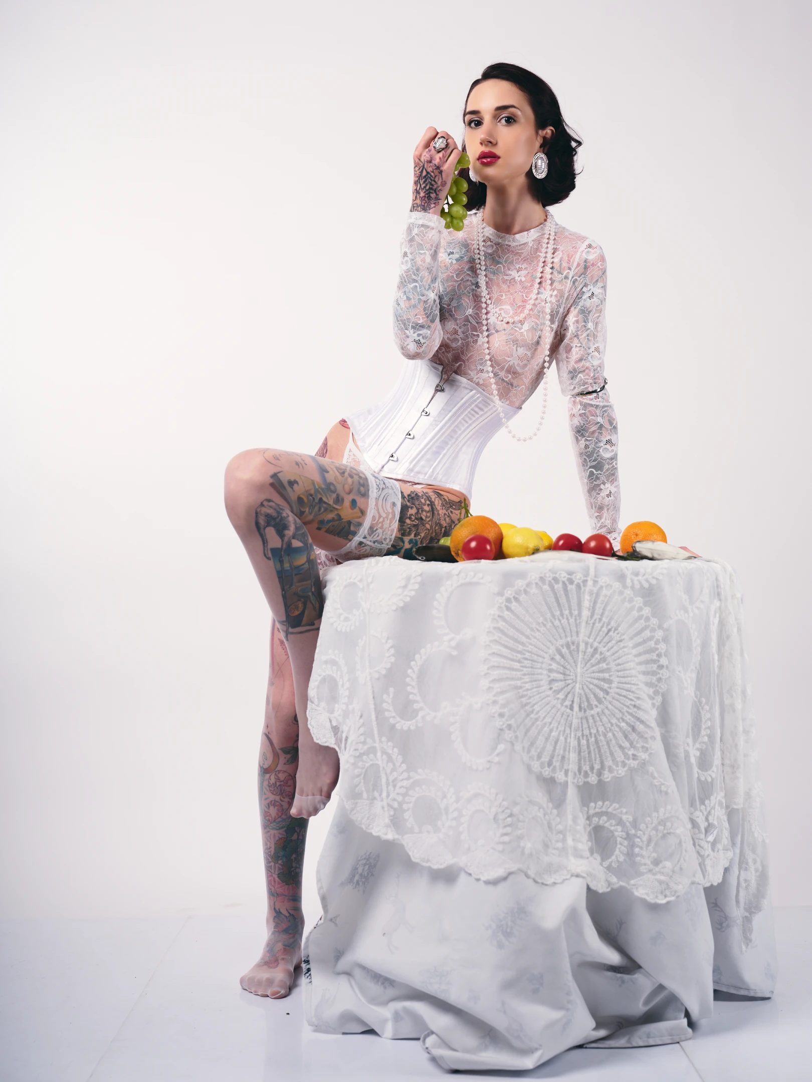 A poised woman with dark, wavy hair styled in a vintage updo sits elegantly atop a draped white table, her gaze fixed directly at the viewer with a confident, slightly sensual expression. Her skin is adorned with intricate, colorful tattoos covering her legs and arms, contrasting sharply with the delicate, semi-transparent lace long-sleeve top she wears, which reveals a white corset beneath. She holds a cluster of green grapes near her lips, adding a touch of playful sophistication, while a long strand of pearls drapes down her neck and a large, ornate earring catches the light. The lighting is soft and even, highlighting her features and the textures of her attire and the lace tablecloth...