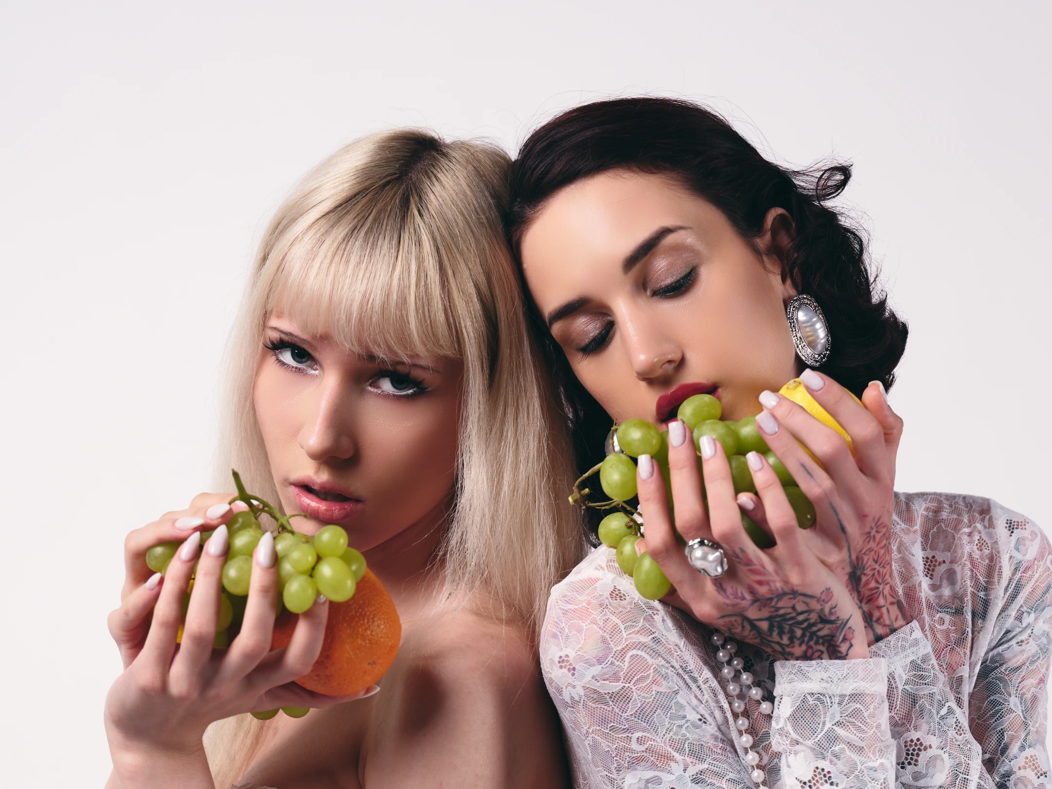 This portrait features two women positioned closely against a stark, neutral white background, emphasizing their contrasting styles and expressions. The woman on the left, with platinum blonde hair and sharp bangs, gazes directly at the viewer with a cool, intense stare, holding a cluster of green grapes and an orange in her hands; her nails are painted a soft white, and she wears minimal makeup, accentuating her piercing eyes. The woman on the right, with dark, wavy hair styled in a vintage updo, closes her eyes as she leans into the frame, savoring a grape cluster while holding a lemon; her attire includes a delicate lace blouse with floral embroidery, adorned with a pearl necklace and ...