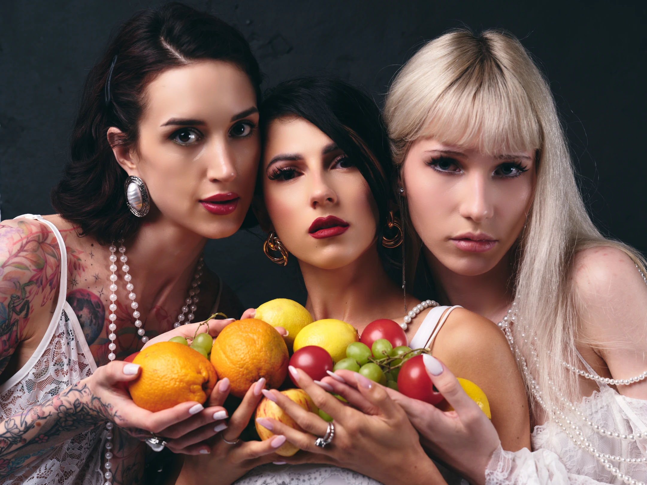 This portrait features three women posing closely together against a dark, textured backdrop that emphasizes their vibrant presence. The woman on the left has dark, wavy hair with a side part, adorned with large, ornate earrings and a pearl necklace; her arms are covered in colorful tattoos, and she wears a delicate white lace top. The center woman has sleek black hair, bold red lipstick, and gold hoop earrings, holding a bowl of fruit with a neutral, confident expression. The woman on the right sports long blonde hair with bangs, wearing a white lace dress with layered pearl necklaces and subtle makeup; she gazes directly at the camera with a serious, composed look. Their hands cradle a ...