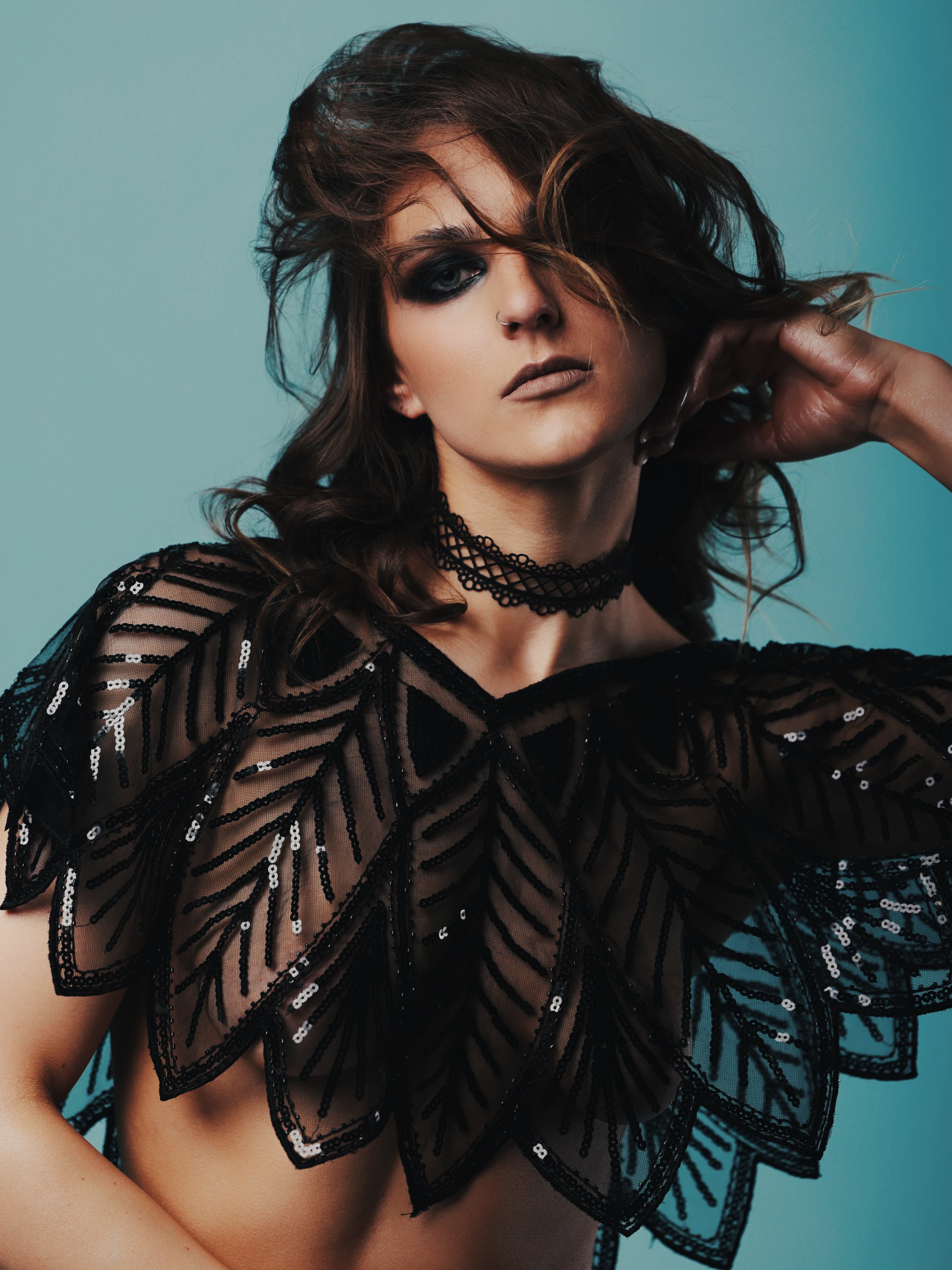 A woman with dark, windswept hair and smoky eye makeup gazes directly at the camera with a poised, intense expression. She wears a dramatic, black, sequined, leaf-patterned capelet that drapes over her shoulders and chest, paired with a matching lace choker. Her hand gently touches her hair, adding a sense of movement and elegance. Soft studio lighting highlights the shimmer of the sequins and the texture of her attire, while the cool teal background creates a moody, high-contrast atmosphere. The image conveys a sense of bold sophistication and artistic drama.