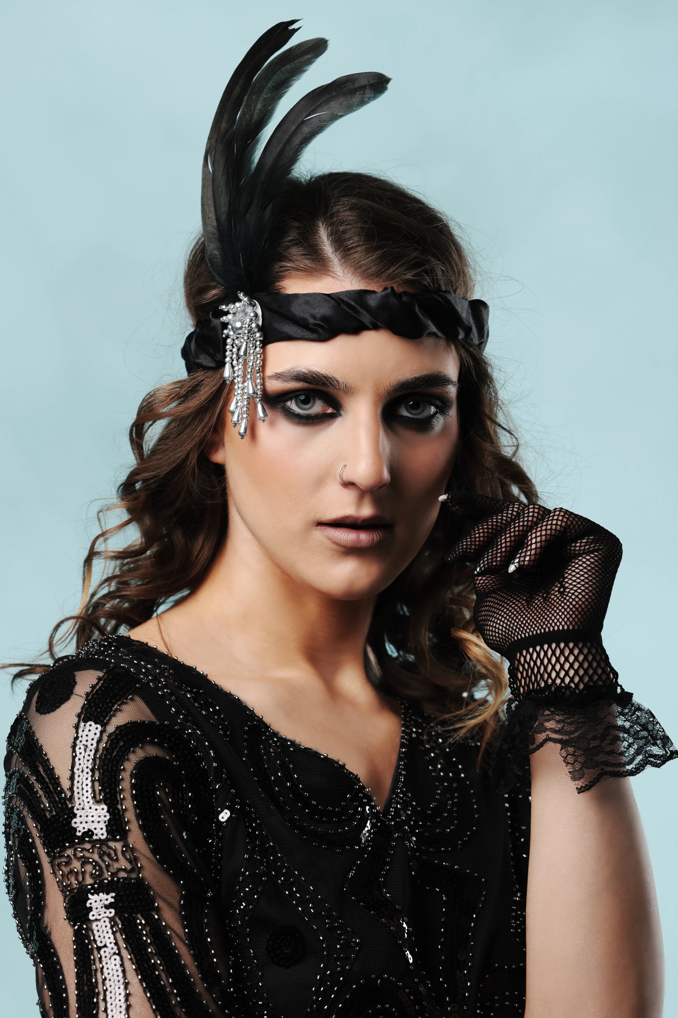 A woman with dark, wavy hair styled in a 1920s flapper look wears a black feathered headpiece and a beaded, sequined black dress with lace sleeves. Her expression is poised and direct, gazing forward with bold, dark eye makeup and a subtle lip color. She holds a black mesh glove near her face, adding to the vintage glamour. Soft studio lighting highlights the sparkle of her attire against a pale, cool-toned background, evoking a sophisticated, retro mood. The image captures elegance and confidence with a strong 1920s flapper aesthetic.