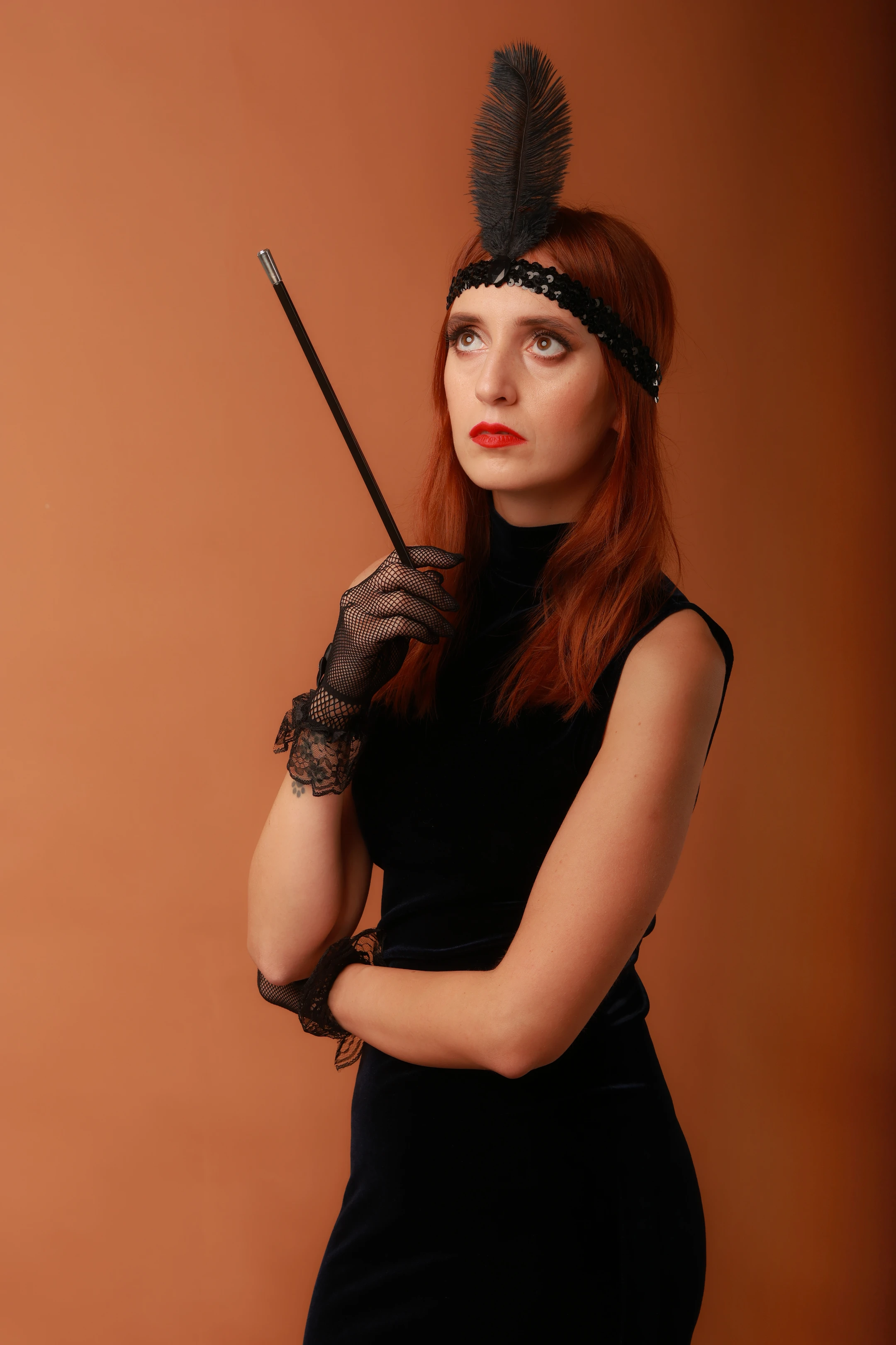 A woman with vibrant red hair stands against a warm, solid orange background, wearing a black velvet dress, black lace gloves, and a black feathered headband. She holds a slender black cane in her right hand, her gaze directed upward and to the side with a poised, contemplative expression. Soft lighting highlights her features and the texture of her attire, evoking a 1920s flapper aesthetic. Her posture is elegant and slightly defiant, with arms crossed and one hand resting on the cane, suggesting confidence and mystery. The overall mood is sophisticated, stylized, and timeless.