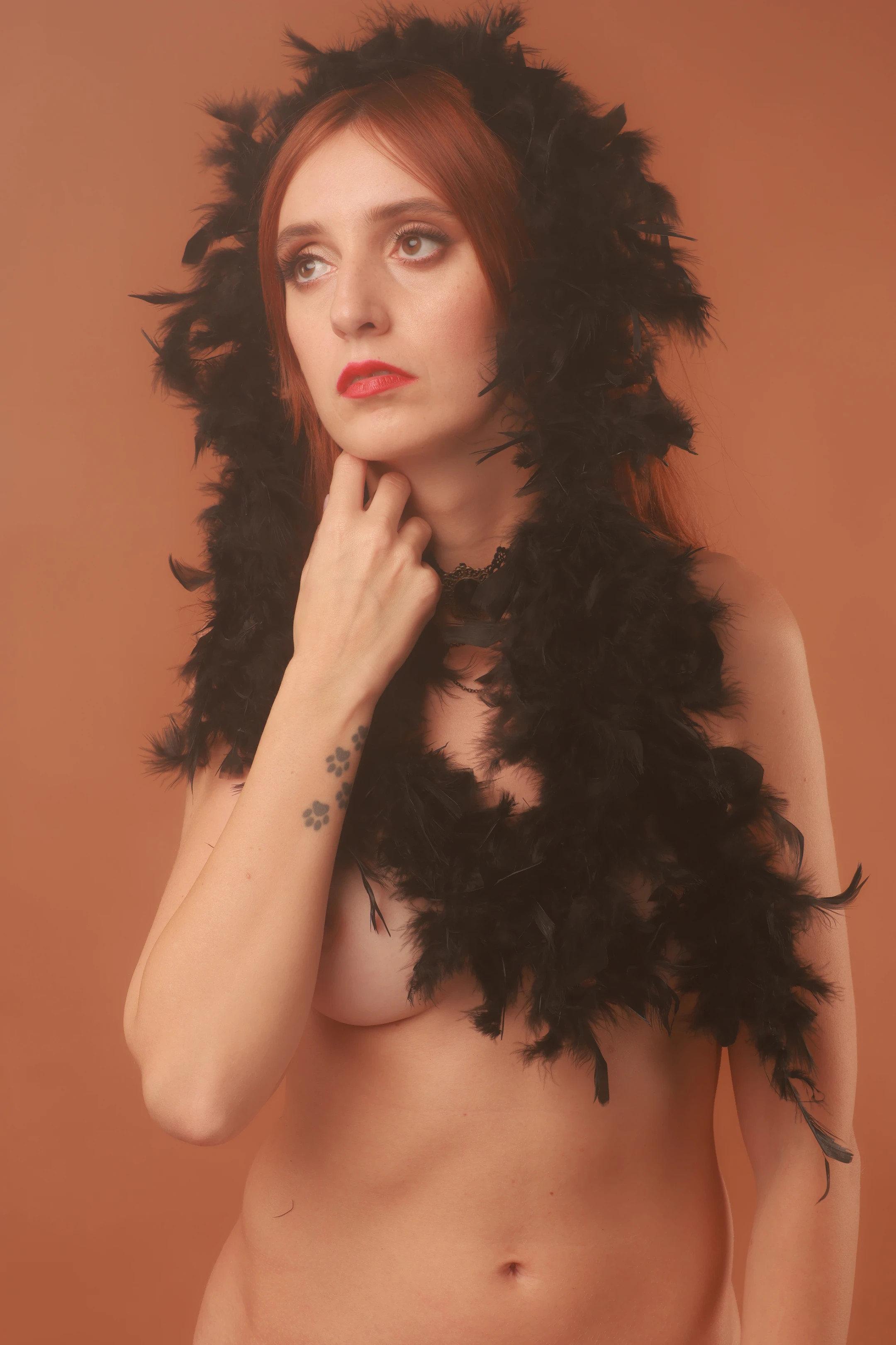 A nude woman with reddish-brown hair and a black feathered headpiece and scarf gazes upward with a contemplative, serene expression, her right hand gently resting near her chin. Her left arm is bent, hand near her neck, while a small floral tattoo adorns her left forearm. Soft, warm lighting bathes her in a muted orange glow, emphasizing the texture of the feathers and the smoothness of her skin. The background is a solid, unbroken orange, creating a minimalist, intimate atmosphere that draws focus to her poised, slightly melancholic demeanor. The composition is centered, evoking a sense of quiet elegance and mystery.