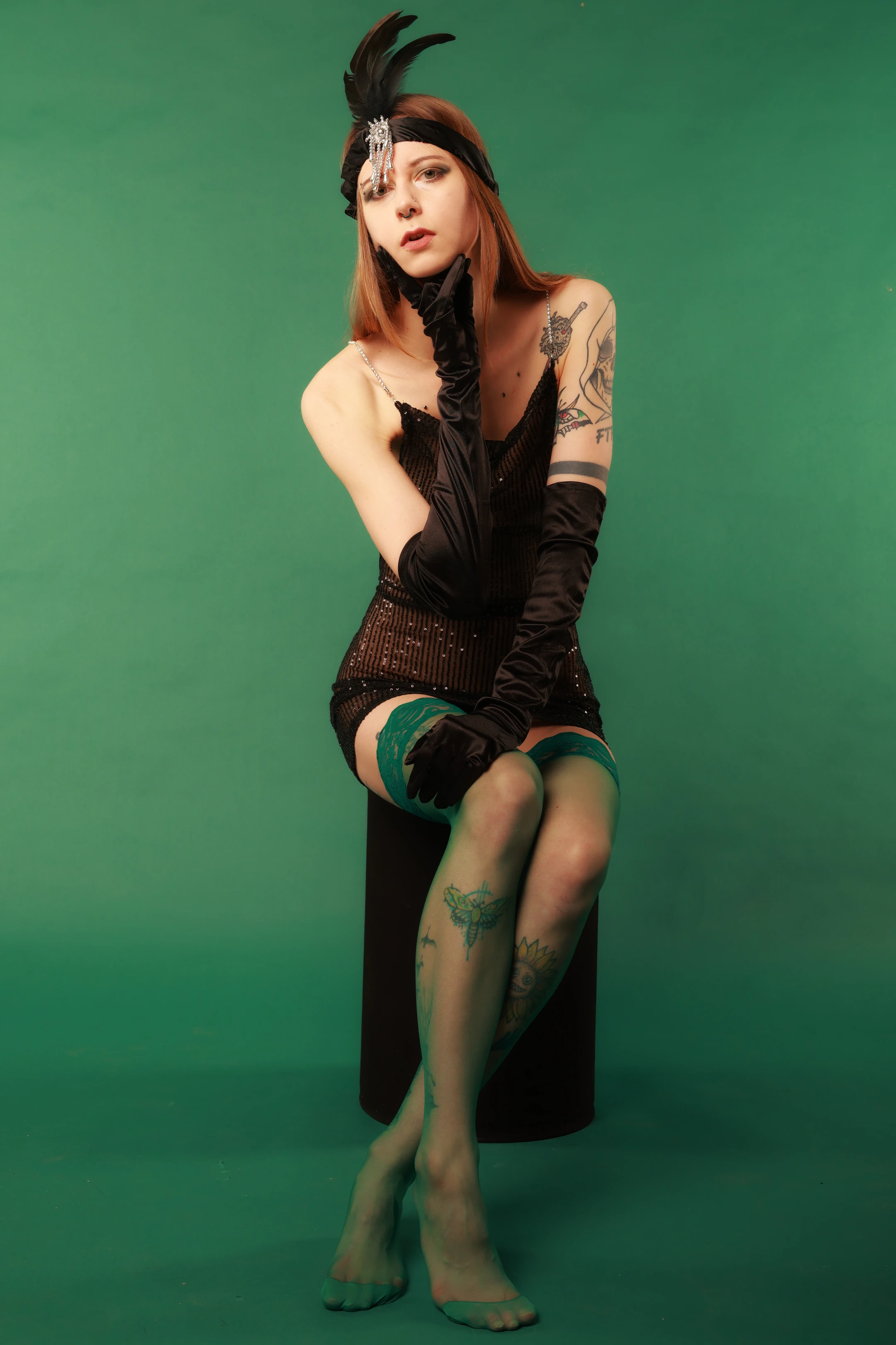 A woman with long, light brown hair sits elegantly against a solid, vibrant green backdrop. She wears a black, beaded, one-shoulder dress with long, sheer gloves and green-tinted stockings, accented by tattoos on her arms and legs. Her head is adorned with a black feathered headpiece and a silver headband, and she gazes directly at the camera with a poised, neutral expression. Soft studio lighting highlights her features and attire, creating a moody, vintage glamour vibe. The green background is uniform and saturated, emphasizing the subject’s dramatic, stylized appearance.