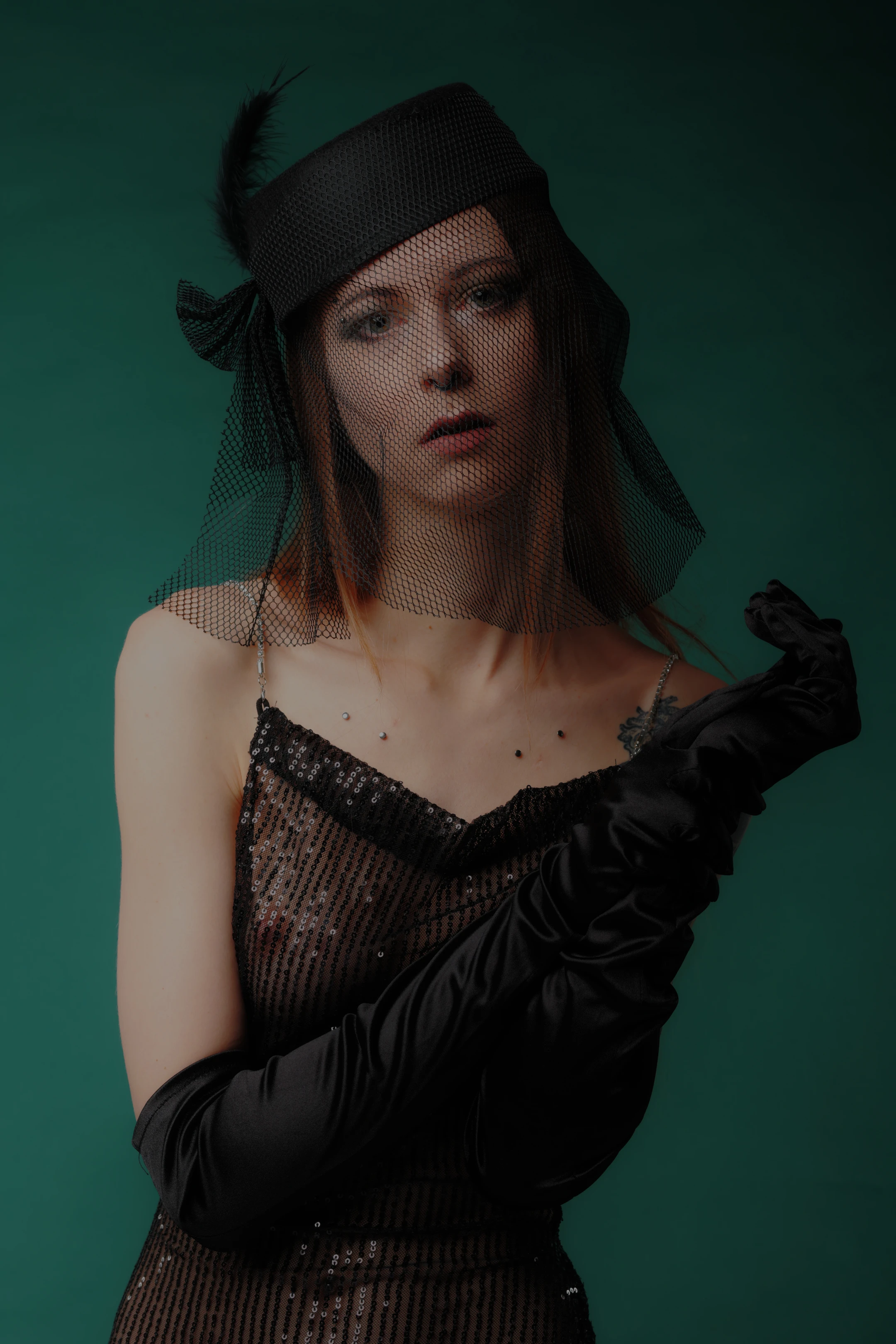 A woman in a vintage 1920s style poses against a solid emerald green backdrop. She wears a black netted veil, a black feathered headpiece, and long black gloves, with a sequined black dress featuring a plunging neckline. Her expression is serious and direct, gazing forward with a slight frown, while one gloved hand rests near her chest and the other near her shoulder. Soft lighting highlights her face and the shimmer of her dress, creating a moody, elegant atmosphere with a hint of mystery. The deep green background contrasts sharply with her dark attire, emphasizing her poised, dramatic presence.
