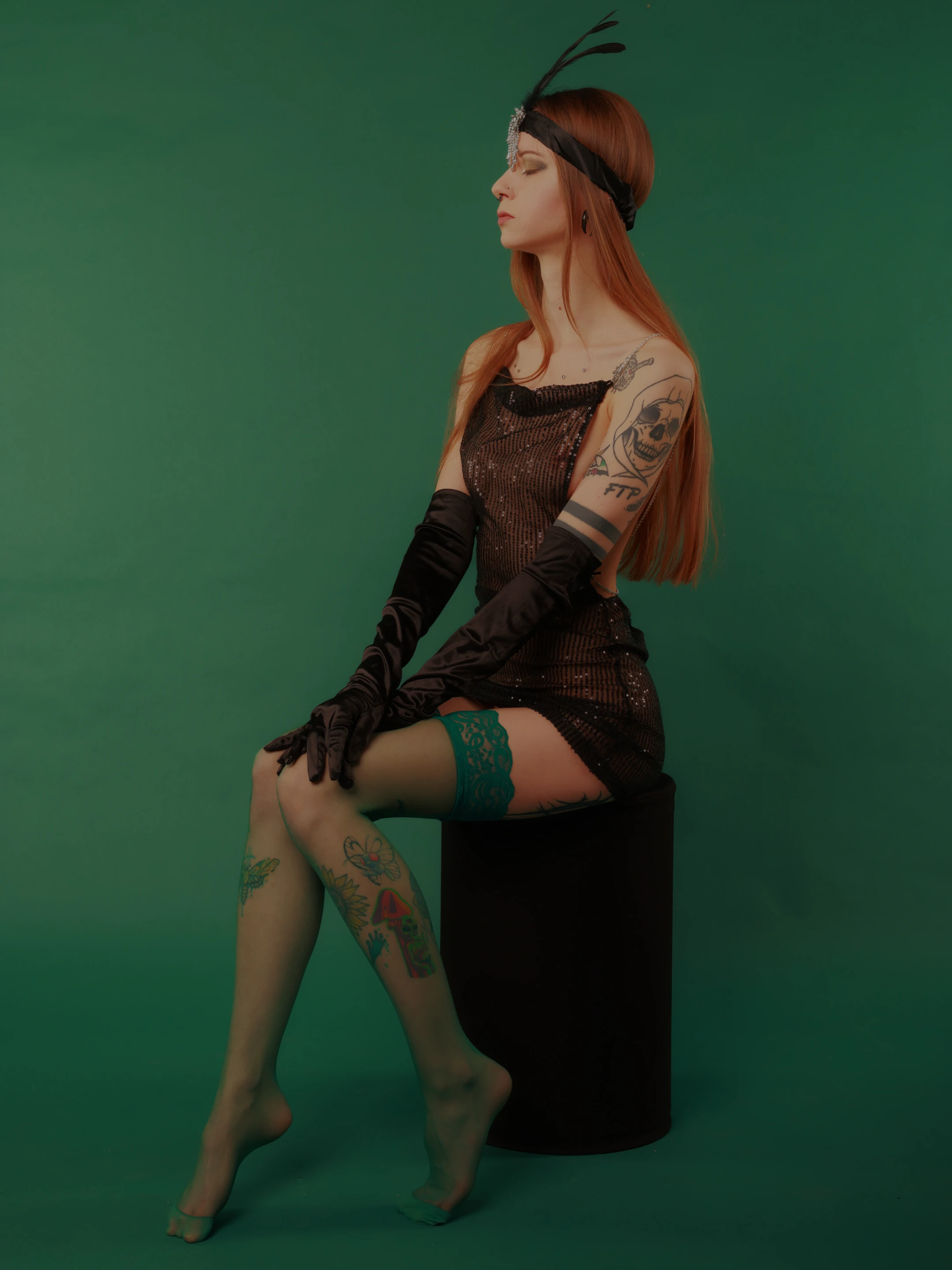 A woman with long, flowing red hair sits elegantly on a black stool against a solid green backdrop. She wears a shimmering black dress, long black gloves, and a feathered headpiece, exuding a vintage, flapper-inspired style. Her expression is serene, with closed eyes and a slight smile, suggesting calm confidence. Soft lighting highlights her tattoos on arms and legs, adding texture and artistry to her appearance. The mood is sophisticated and timeless, with the green background creating a bold, monochromatic contrast to her dark attire and vibrant hair.