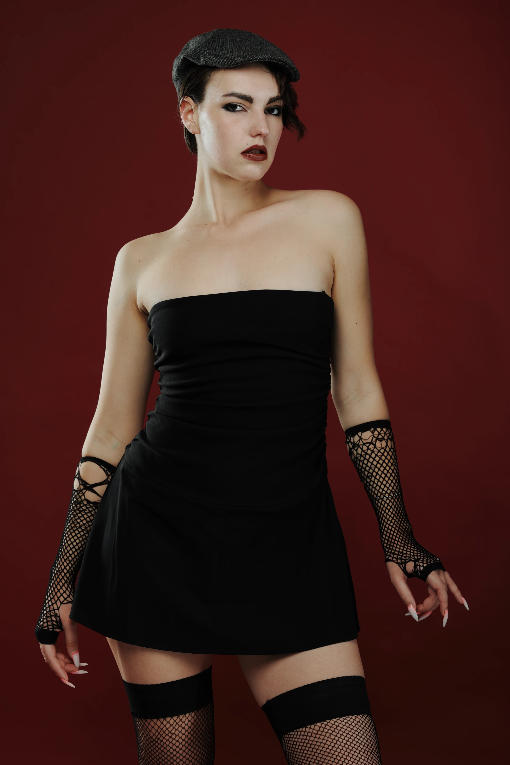 A woman with dark, styled hair and bold makeup poses confidently against a deep red background. She wears a black strapless mini-dress, fishnet gloves, and fishnet stockings with black garters, complemented by a black beret. Her expression is intense and direct, with a slight tilt of the head and a firm gaze toward the camera. Soft studio lighting highlights her features and outfit, creating a moody, edgy aesthetic. The rich red backdrop enhances the dramatic, vintage-inspired fashion look, emphasizing her poised, assertive presence.