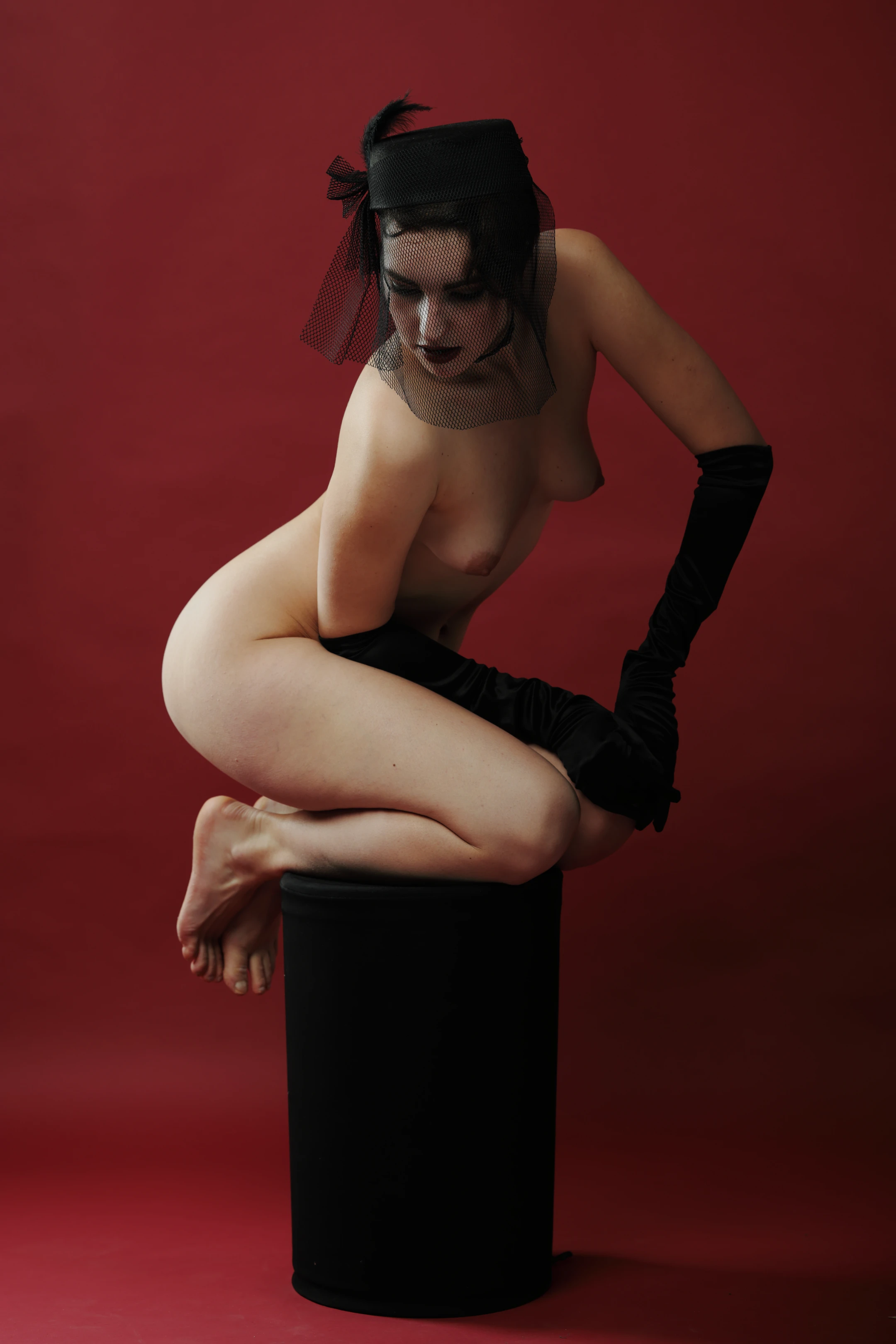A nude woman sits atop a black cylindrical stool against a deep red backdrop. Her dark hair is partially covered by a black headpiece and a sheer black veil, with one long black glove adorning her right hand resting on her thigh. Her left hand is bare, resting near her knee, and she gazes slightly downward with a neutral, contemplative expression. Soft lighting highlights her curves while casting subtle shadows, creating a moody, elegant atmosphere. The stark red background contrasts with her black accessories, emphasizing her form and the surreal, stylized composition.