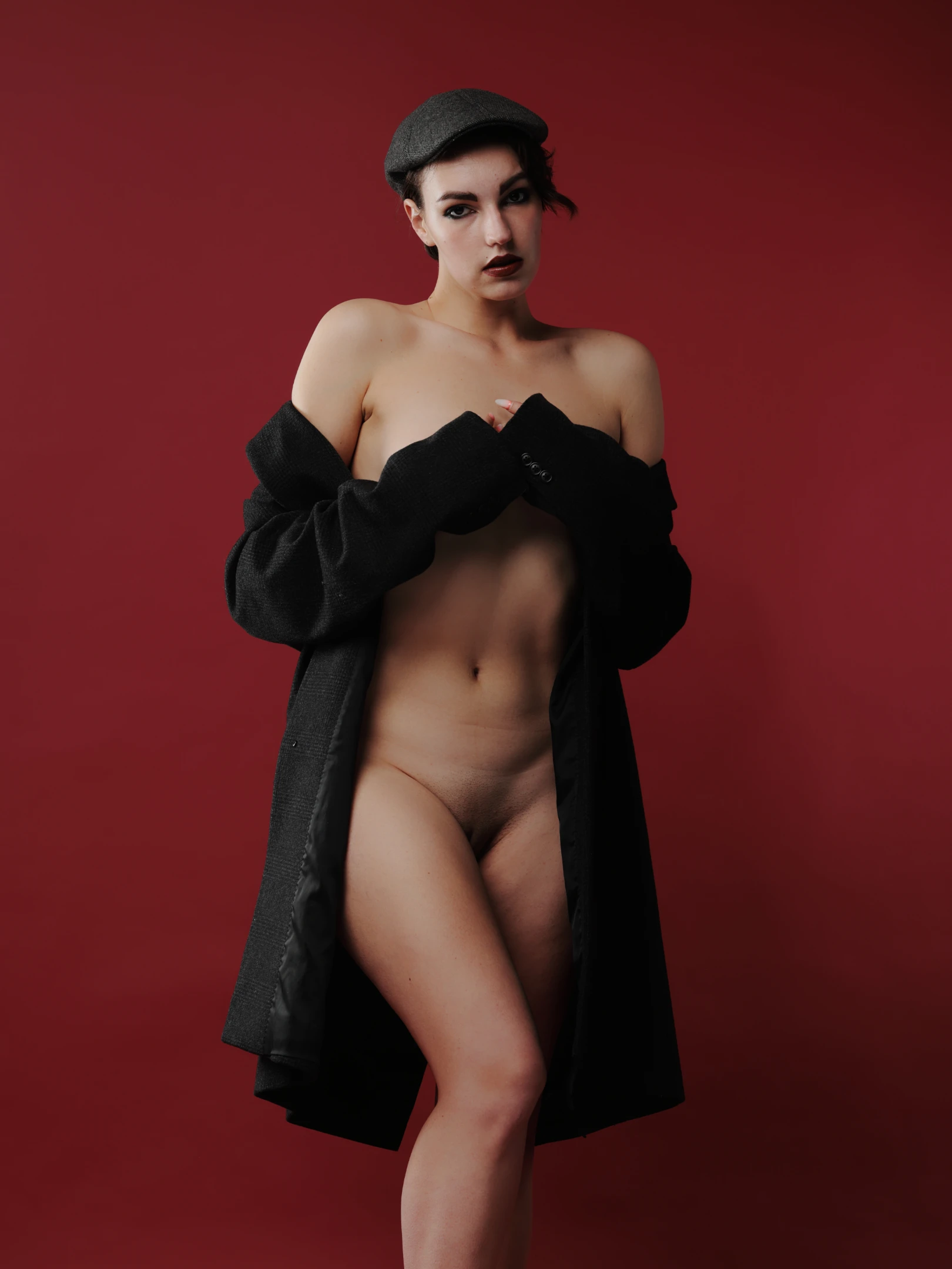 A woman with short dark hair and a dark beret stands against a deep red background, her expression neutral and direct, gazing forward. She is nude except for a long, loose black coat draped over her shoulders and arms, which she holds open with both hands, revealing her body. The lighting is soft and focused, highlighting her form against the rich, saturated red backdrop. The mood is bold and sensual, with a strong contrast between the black garment and the vibrant background, emphasizing her poised, confident stance.