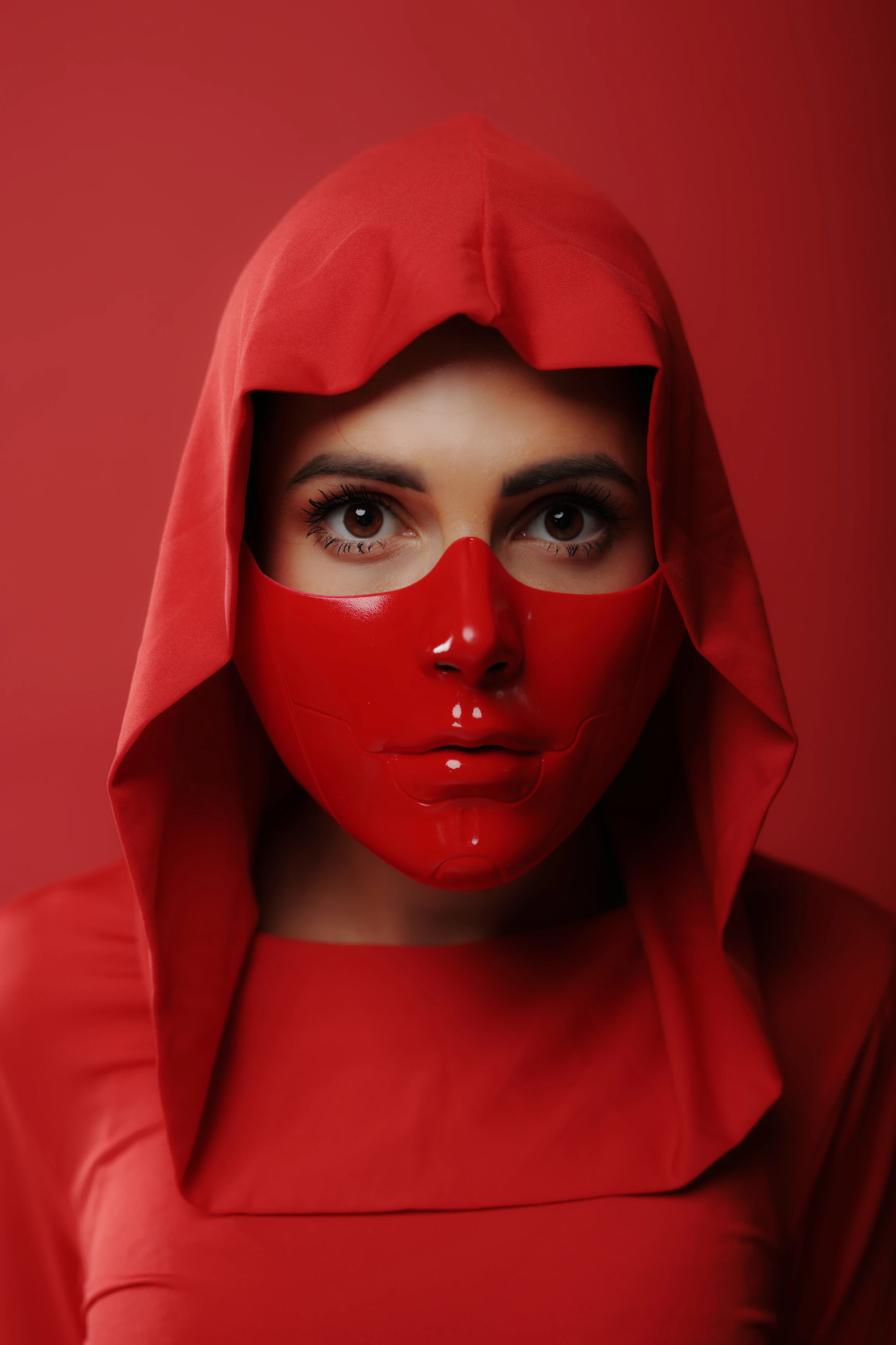 A woman with fair skin and dark, expressive eyes gazes directly at the viewer, her face partially obscured by a glossy, red, form-fitting mask that covers her nose, mouth, and chin. She wears a matching red hooded garment that drapes over her shoulders and head, creating a seamless, monochromatic look. The lighting is soft yet dramatic, highlighting the mask's sheen and the subtle contours of her face beneath. The background is a solid, deep red, enhancing the portrait's intense, mysterious mood. Her expression is neutral yet compelling, evoking intrigue and power through the stark, unified red palette.