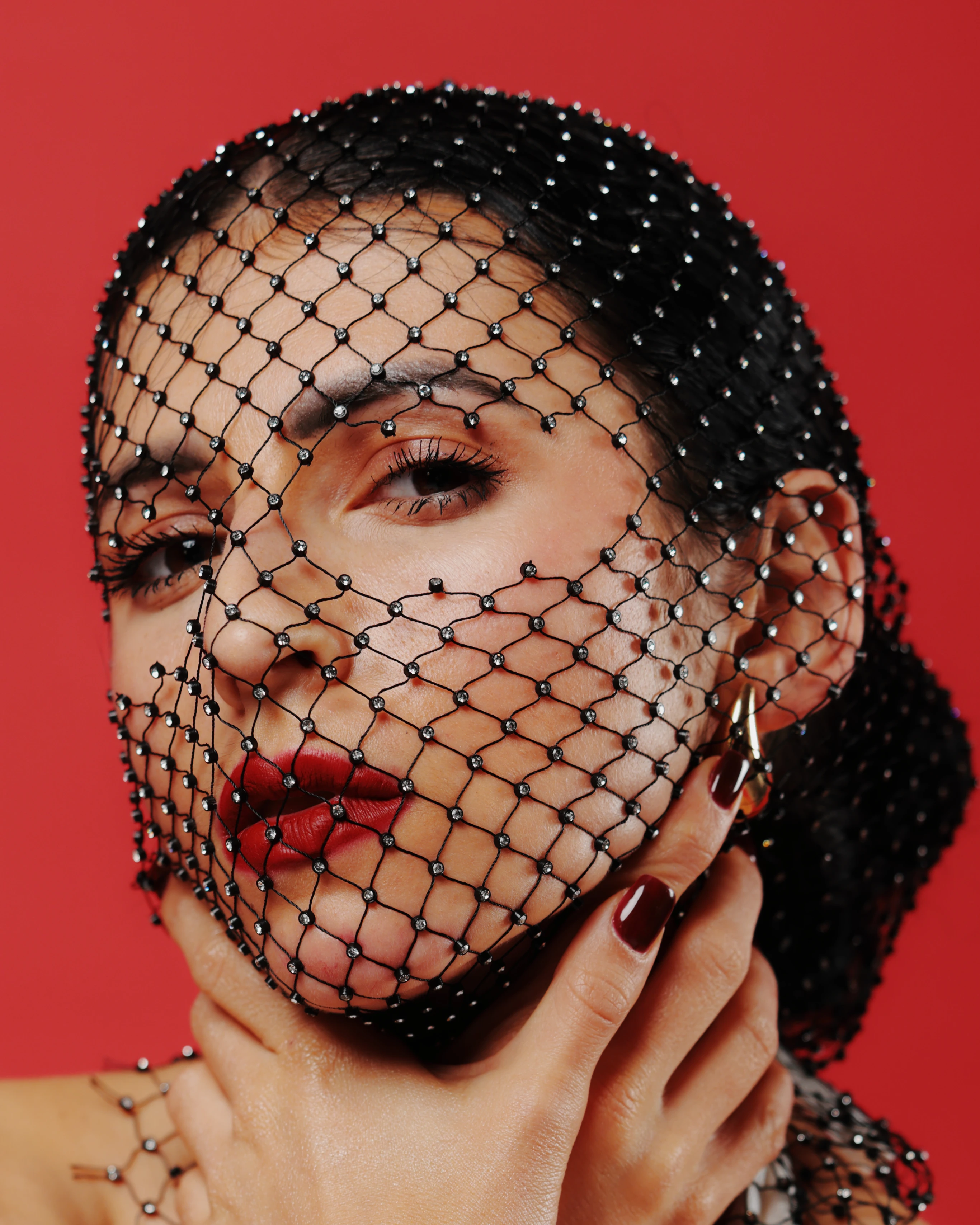 A woman with dark, glossy hair pulled back in a sleek updo gazes directly at the viewer, her face partially obscured by a black beaded net veil that drapes over her head and shoulders. Her lips are painted a bold red, contrasting with her dark, polished nails and a delicate gold hoop earring. She holds the veil gently with both hands, fingers lightly touching the fabric near her jawline, conveying a poised, mysterious expression. The lighting is soft yet dramatic, highlighting her features against a vibrant, solid red background that amplifies the intensity and elegance of the portrait. The mood is sensual and enigmatic, with a touch of theatrical sophistication.