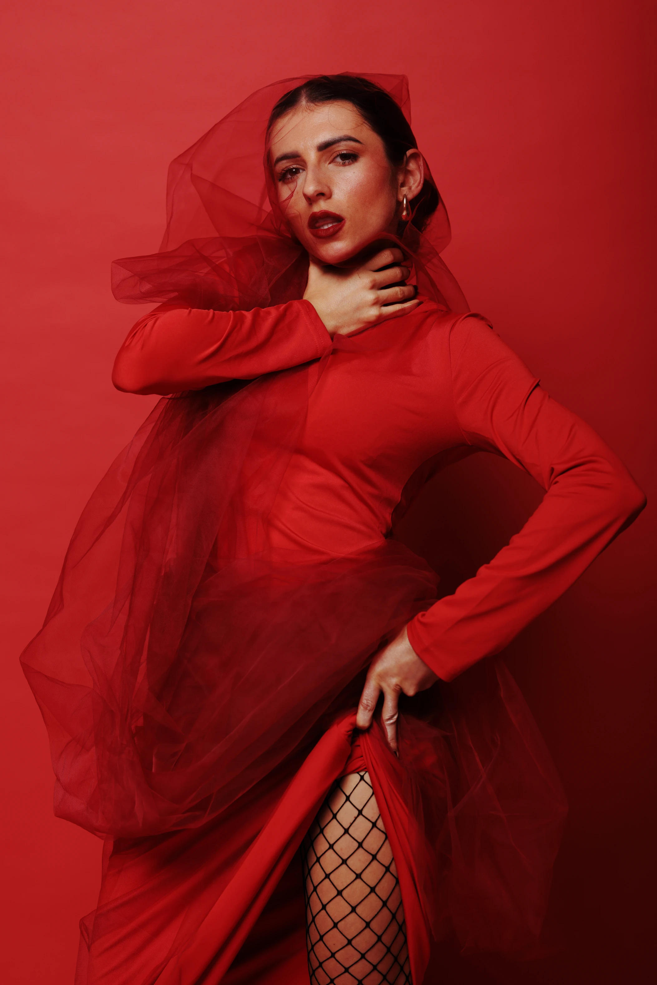 A woman with dark hair and bold red lipstick stands against a solid red background, her expression confident and slightly defiant. She wears a form-fitting red long-sleeve dress layered with a sheer, flowing red veil that drapes over her head and cascades down her body, partially revealing fishnet stockings. Her pose is dynamic—hands on her neck and hip—suggesting movement or drama. Lighting is soft yet directional, highlighting her features and the texture of the fabric. The mood is bold, sensual, and theatrical, with the monochromatic red palette creating a powerful, unified visual impact.