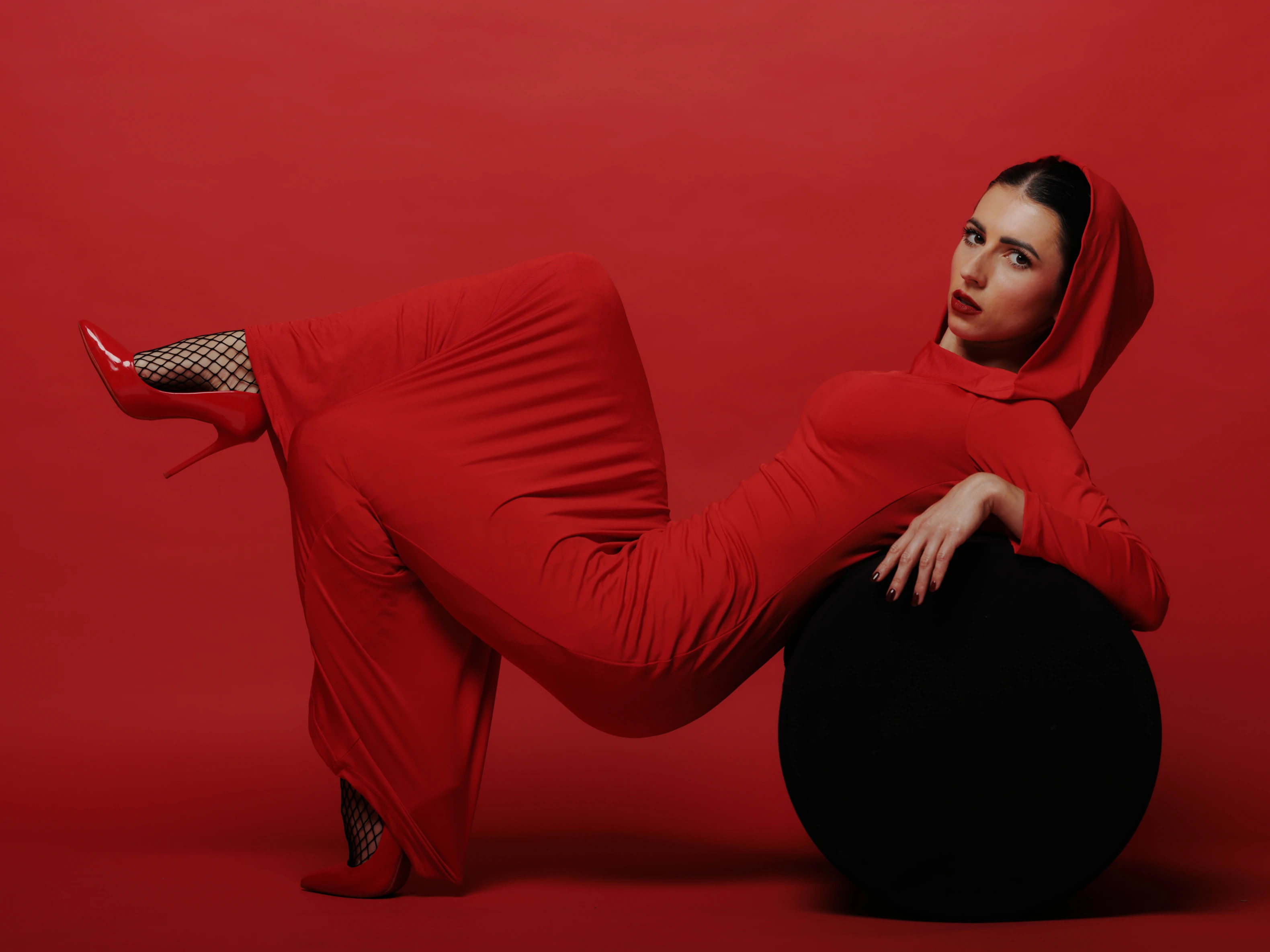 A woman in a full-length red hooded dress reclines on a black spherical object, one leg bent and the other extended, wearing red high-heeled shoes and black fishnet stockings. Her dark hair is pulled back, and she gazes directly at the camera with a neutral, poised expression. The lighting is even and studio-like, emphasizing the vibrant red tones of her outfit and the stark contrast with the black sphere and background. The solid red backdrop creates a bold, minimalist mood, focusing attention on the subject's form and the dramatic interplay of color and texture. Her pose conveys confidence and stylized elegance, with one hand resting on the sphere and the other near her hip.