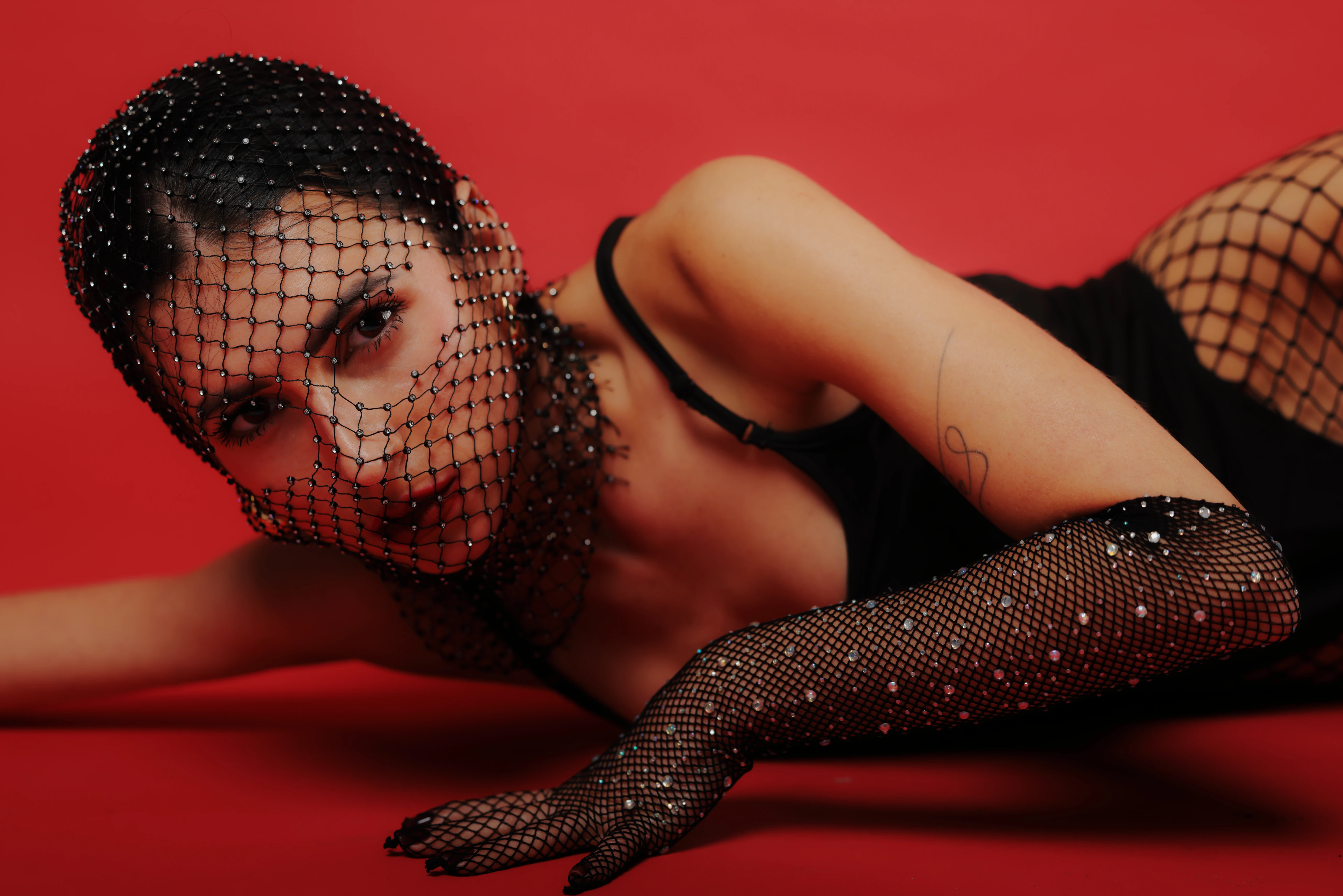 A woman lies on a vibrant red background, wearing a black netted face veil and matching fishnet gloves adorned with small white dots. Her dark hair is pulled back, and she gazes directly at the camera with a neutral, intense expression. A subtle tattoo is visible on her left forearm. The lighting is dramatic, highlighting her features and the texture of the netting, creating a sultry, high-contrast mood. The bold red background emphasizes the black elements, evoking a sense of glamour and mystery.