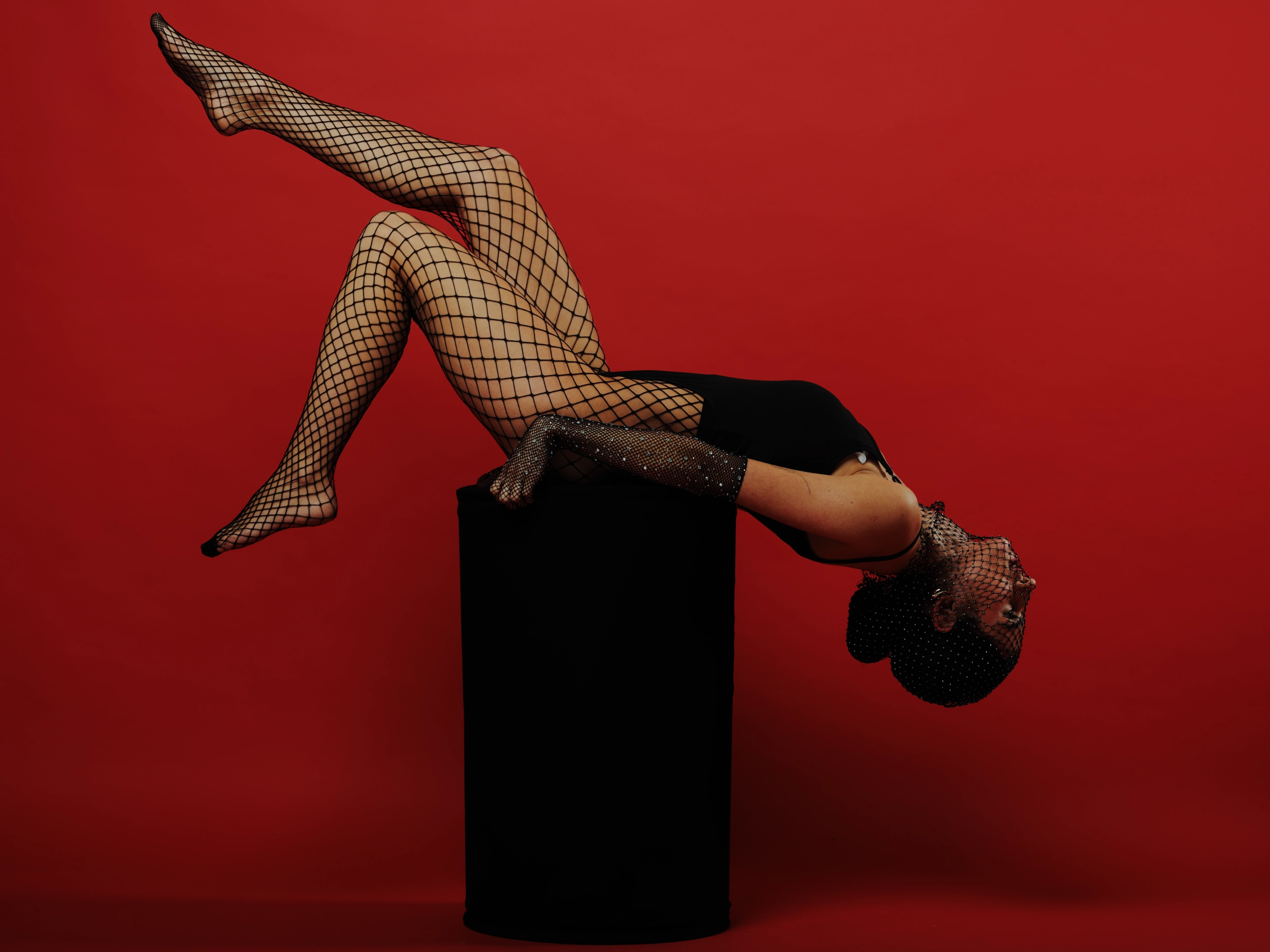 A woman in a black bodysuit and fishnet stockings, with a matching fishnet headpiece, is inverted, balancing on a black cylindrical pedestal. Her legs are extended upward, toes pointed, while her head rests on the cylinder’s top, face turned away from the camera. The lighting is studio-style, highlighting her form against a solid, vibrant red background. The mood is bold and theatrical, emphasizing movement and control. Her expression is neutral, focused, and poised, suggesting performance or artistry. The composition is dynamic, with strong lines and a high-contrast color scheme that draws attention to her body and the stark geometry of the pedestal.