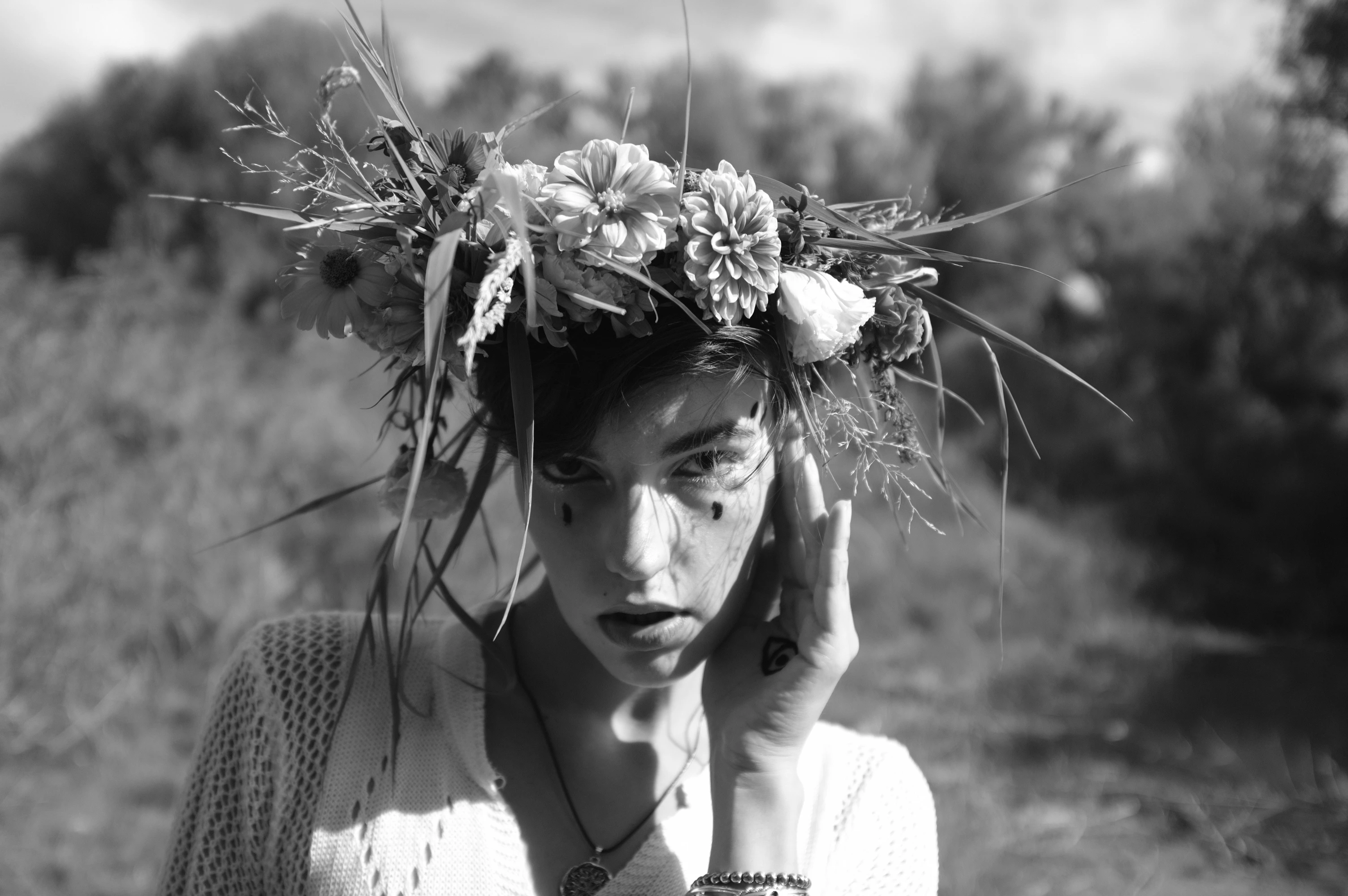 A black-and-white portrait features a person with short dark hair adorned with a large, textured floral crown of mixed flowers and tall grasses. Their face is partially obscured by the crown and they gently touch their cheek and temple with one hand, conveying a contemplative or serene expression. They wear a light, knitted sweater with a visible pendant necklace and a dark, possibly tribal, dot pattern on their cheek. Soft, diffused lighting creates a dreamy, ethereal mood, with the blurred background suggesting an open, natural field under a bright sky. The image evokes a sense of quiet introspection, nature's embrace, and artistic stillness.