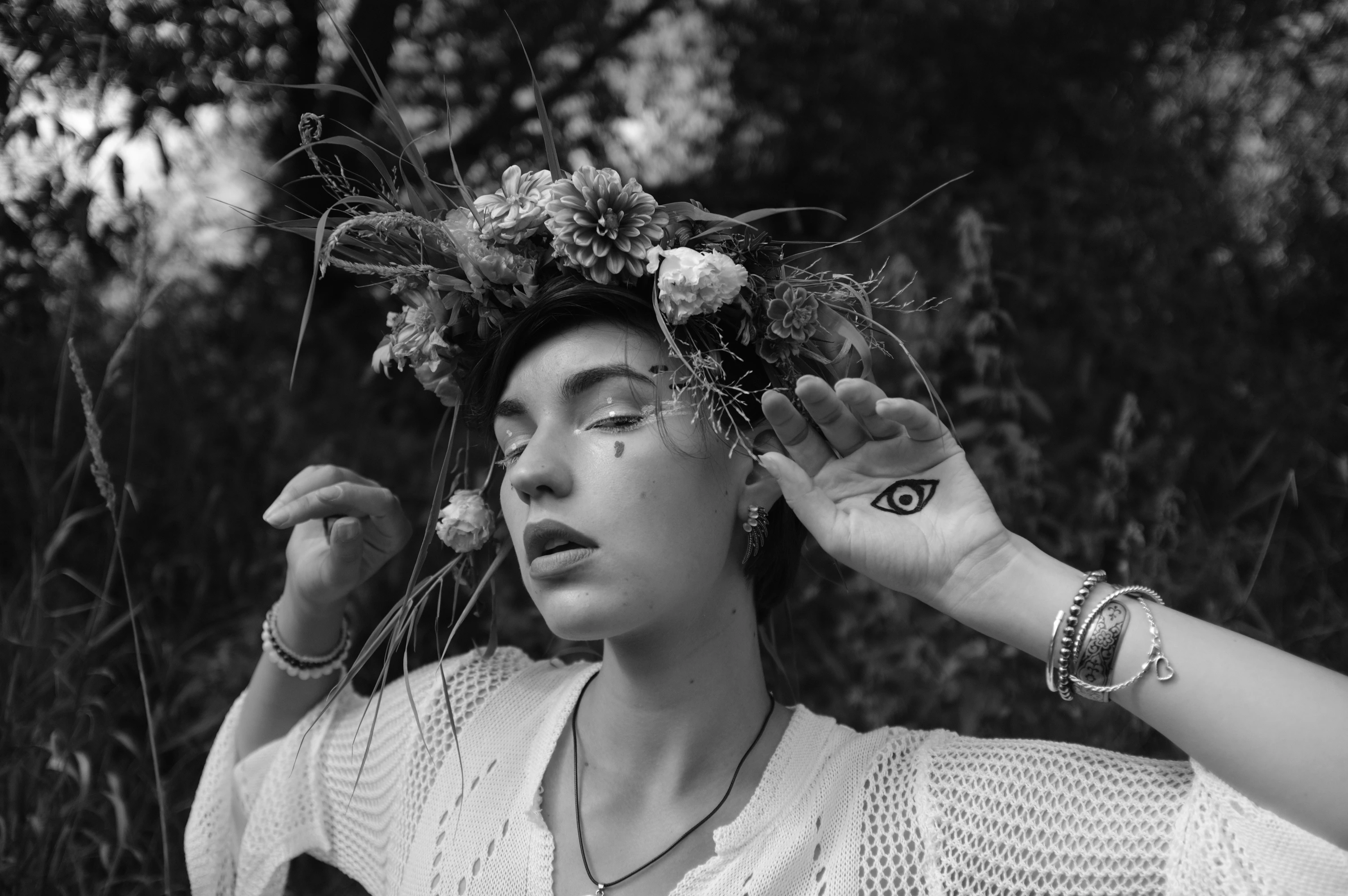 A black-and-white portrait captures a woman with a serene, contemplative expression, eyes gently closed or half-lidded, as if in quiet meditation or sunlight. Her head is crowned with a lush, textured floral and grassy wreath, and a dark eye tattoo adorns her left hand, which she raises toward her face. She wears a light, open-weave knit top with a simple necklace, and her right hand rests near her cheek, fingers slightly curled. The lighting is soft and diffused, suggesting natural daylight, with a blurred, leafy background that enhances the ethereal, nature-infused mood. The image conveys tranquility, mysticism, and a deep connection with the natural world.