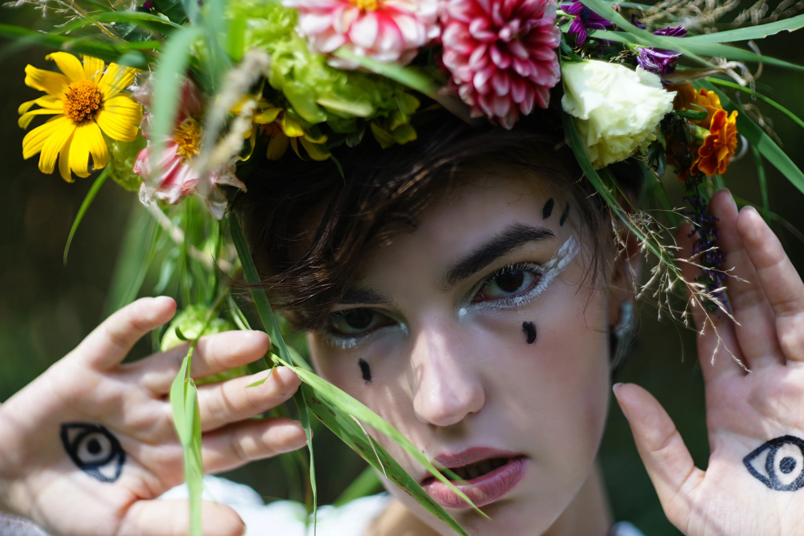 A young person with dark, tousled hair wears a vibrant, overflowing floral crown of pink, yellow, and white blooms, accented with greenery. Their face features bold white eyeliner and three black dots—two on cheeks, one on the nose—while their hands, adorned with black eye tattoos, frame the face with delicate, leafy fingers. They gaze directly at the camera with a neutral, intense expression, mouth slightly open. Soft, natural lighting highlights their features, creating a dreamy, earthy mood. The background is blurred greenery, emphasizing the floral headpiece and the subject’s surreal, nature-infused aesthetic.