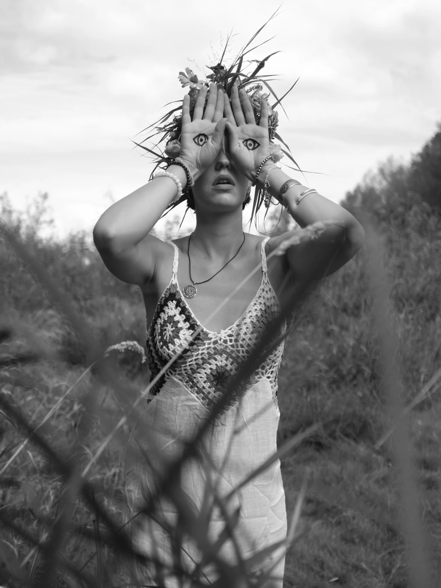 A black-and-white portrait captures a woman standing in tall, wild grass, her face partially obscured by her hands, which are raised to cover her eyes. Her hands are adorned with multiple bracelets, and her face, visible above, shows a startled or curious expression with wide eyes. She wears a textured, crocheted top with thin straps and a simple pendant necklace. The lighting is soft and diffused, suggesting an overcast day, creating a mysterious, ethereal mood. Behind her, blurred vegetation and a pale sky frame the scene, emphasizing her isolation and the surreal, almost mythic quality of her pose. The image's grainy texture and monochrome palette enhance its dreamlike, symbolic atmosp...