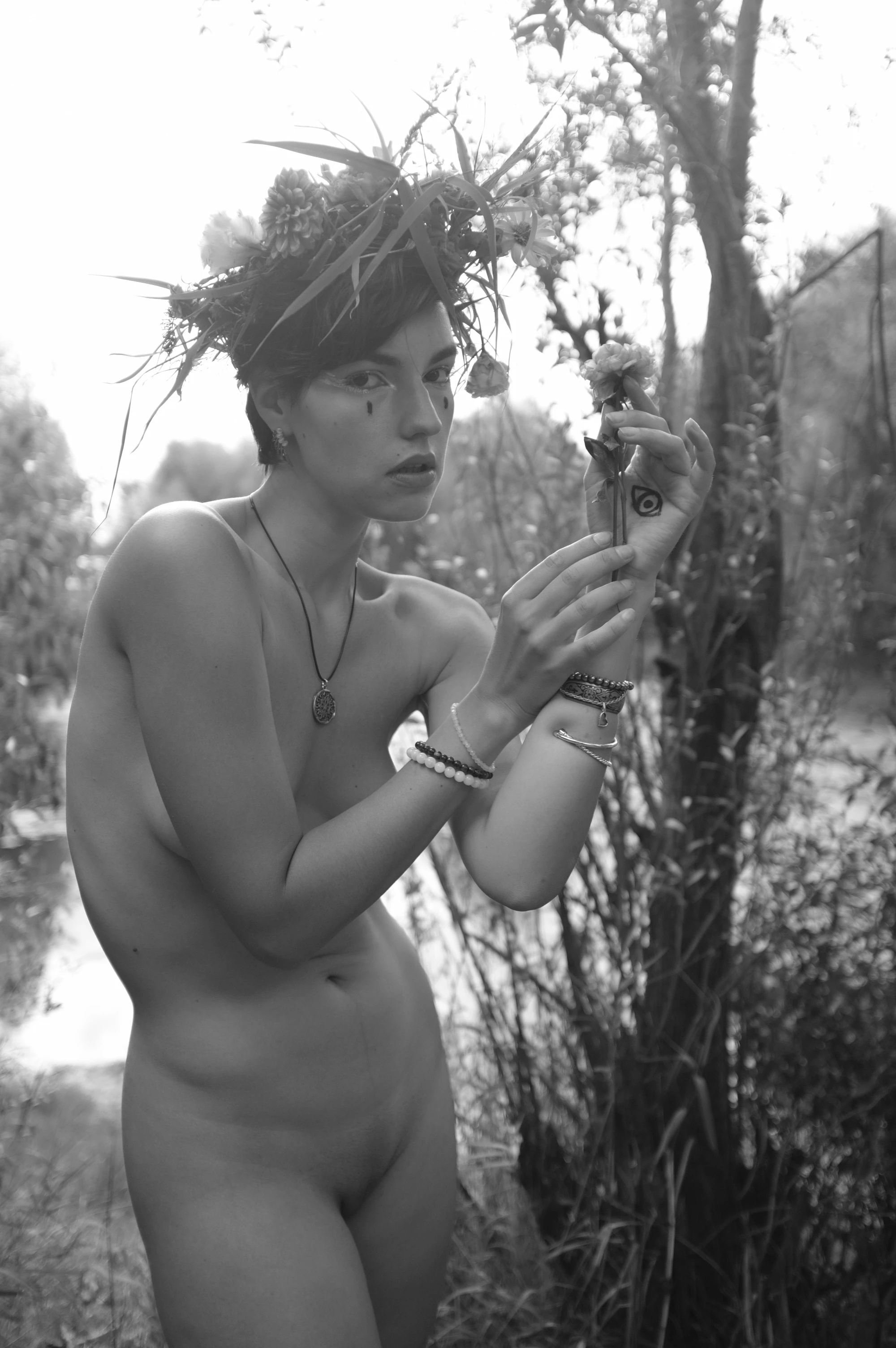 A black-and-white portrait captures a nude woman standing outdoors in tall grass and reeds, her body angled slightly toward the viewer. She wears a crown of tall, spiky leaves and multiple bracelets on her right wrist, with a simple pendant necklace. Her expression is contemplative, gazing downward at a small flower she holds in her left hand, while her right hand rests near her hip. Soft, diffused lighting from above creates a serene, ethereal mood, with the background blurred into a natural, wooded setting. The image conveys quiet introspection and a connection to nature, emphasizing the subject's bareness and the wild, organic elements surrounding her.