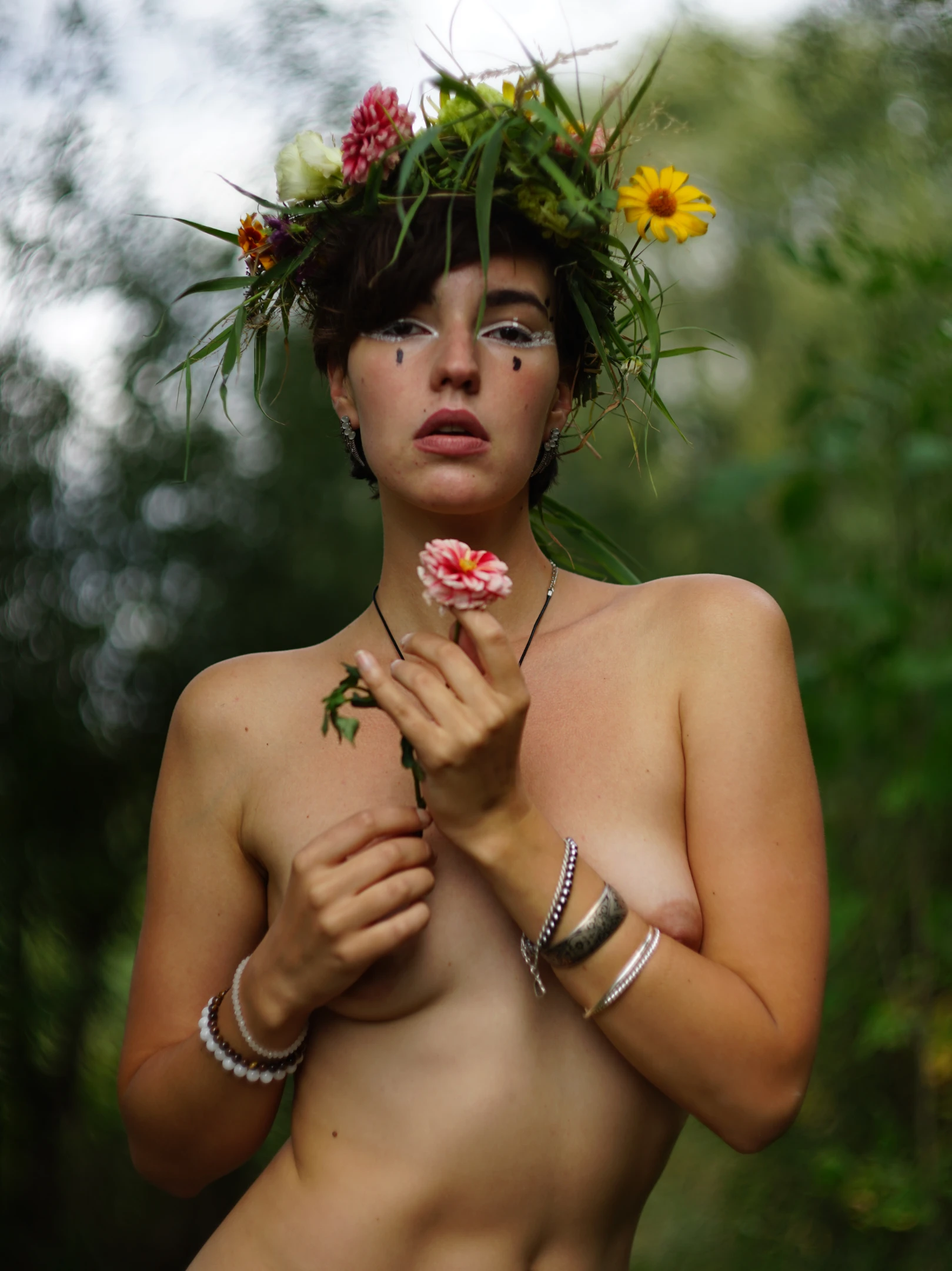 A nude woman stands center-frame, adorned with a lush, vibrant floral crown of green leaves, yellow daisies, and pink blossoms. Her expression is serene yet intense, with dark, smudged eye makeup and a single black dot beneath her left eye, gazing directly at the viewer. She holds a small pink flower in her right hand, while her left hand gently touches her chest, and wears multiple silver-toned bracelets on both wrists. Soft, diffused natural light illuminates her skin, creating a tranquil, earthy mood. The background is a blurred, verdant forest, enhancing the organic, nature-immersed feel of the portrait.