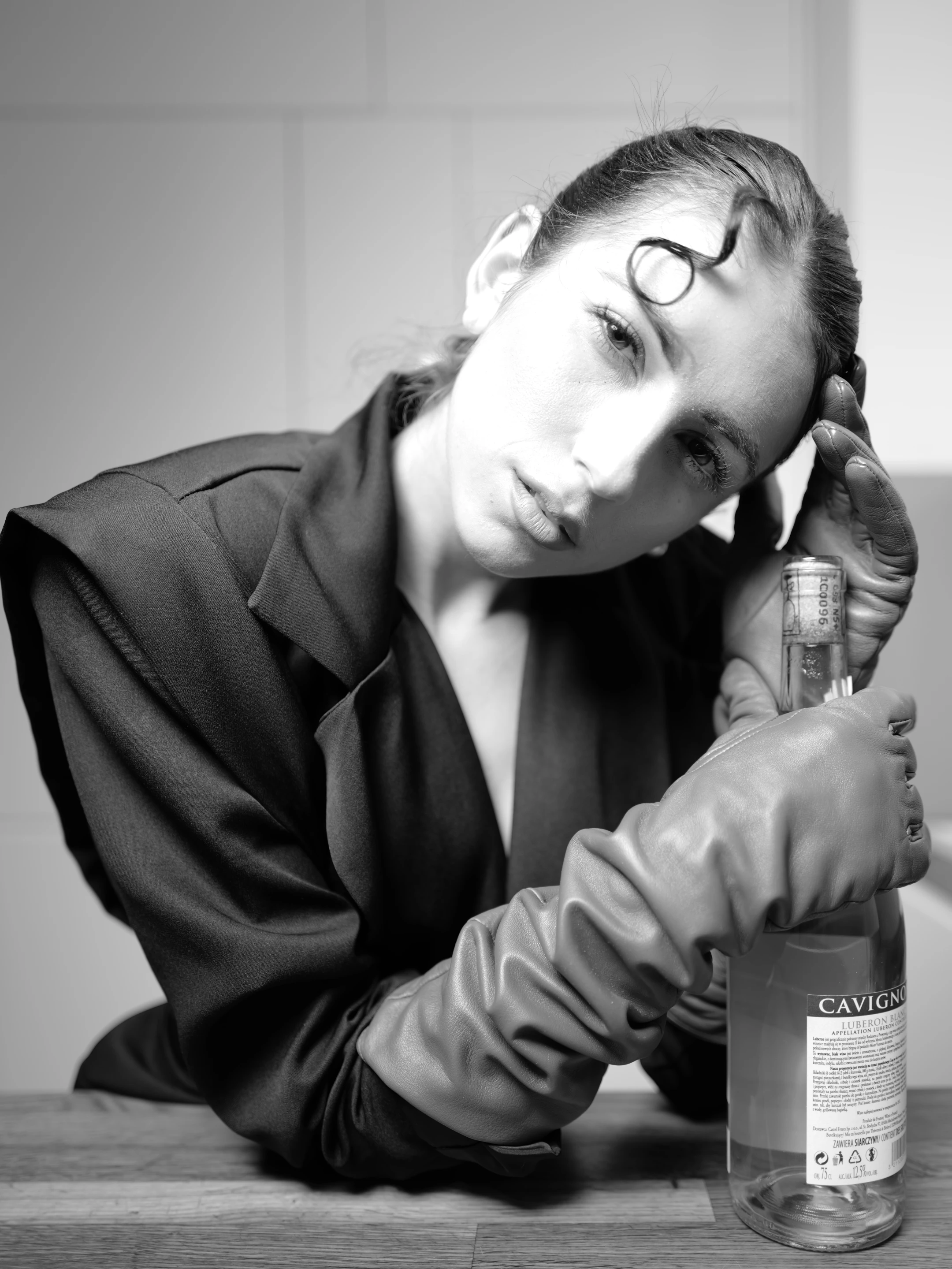 In this striking black-and-white portrait, a woman with dark, wavy hair styled in a modern, slightly tousled updo gazes directly at the camera with a composed, contemplative expression. Her head rests gently against a large, clear bottle of CAVIGNO LUBERON BLANC, a French spirit, while her gloved hands cradle the bottle's base and neck, suggesting a deliberate, almost ritualistic interaction with the object. She wears a sleek, dark blazer with wide, slightly flared sleeves, paired with matching leather gloves that add a tactile, luxurious contrast to the minimalist setting. The lighting is soft yet directional, casting subtle shadows that accentuate her facial features.