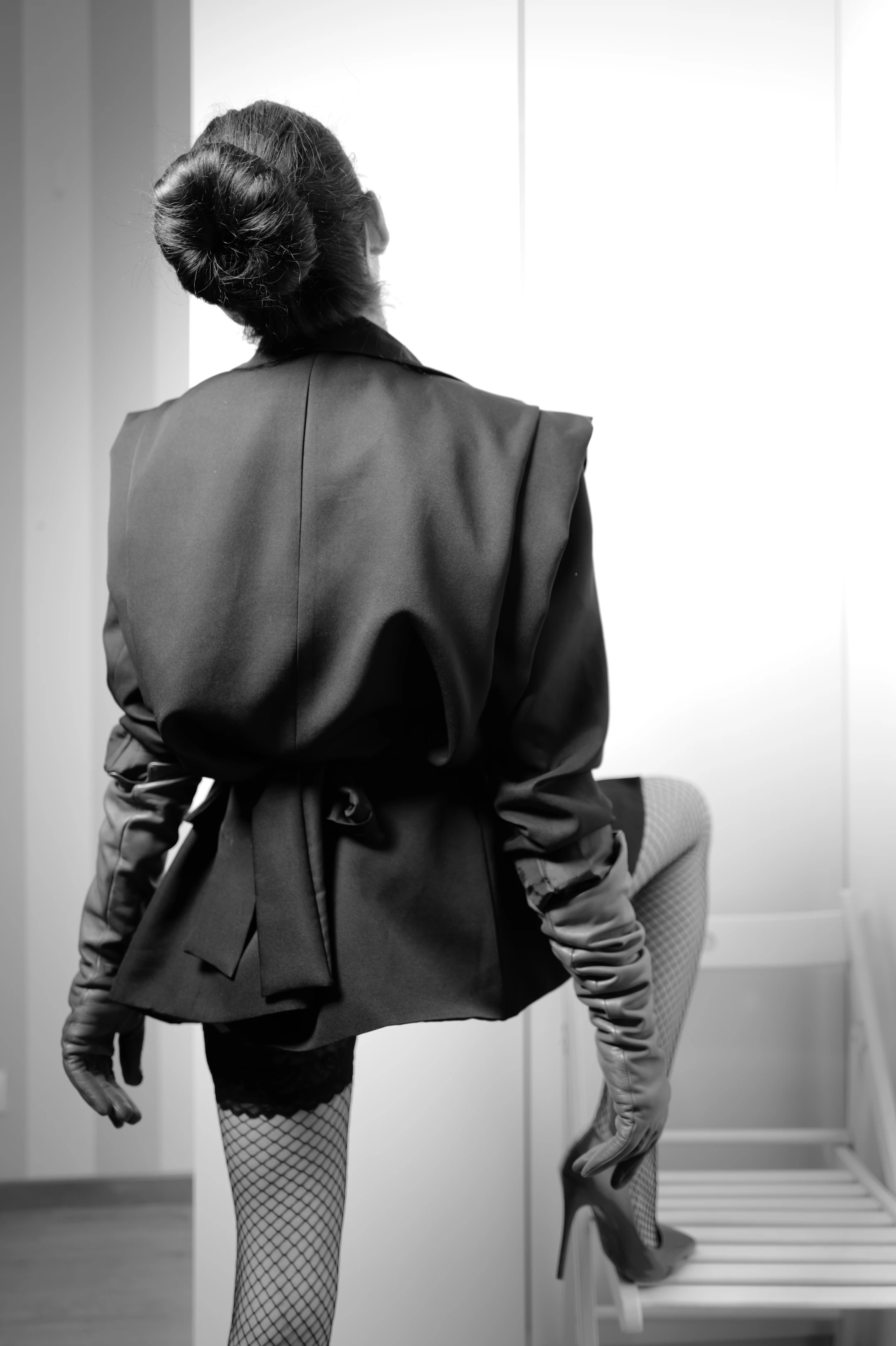This is a striking black-and-white portrait of a woman viewed from behind, seated on a simple white wooden chair, her posture poised and deliberate. Her dark hair is elegantly gathered into a high, voluminous bun, accentuating the sleek lines of her silhouette. She wears a dramatic, oversized black blazer with a cinched waist and a bow detail, paired with fishnet stockings and a lace-trimmed mini skirt, suggesting a blend of power and sensuality. Her long, form-fitting leather gloves extend to her elbows, adding an element of boldness and mystery, while one high-heeled stiletto rests on the chair's edge.