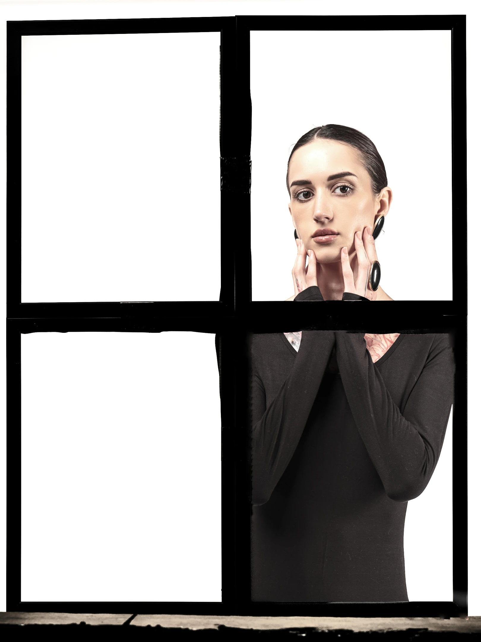 The portrait captures a woman framed within a stark, black-framed window divided into four panes, with the top-right pane containing her face and the bottom-right pane showing her torso. Her dark hair is pulled back neatly, emphasizing her expressive eyes and the subtle makeup that highlights her natural features. She gazes directly at the viewer with a calm, introspective expression, her hands gently cradling her cheeks, fingers delicately placed near her jawline and ears. Her attire consists of a sleek, black long-sleeved top, revealing a hint of tattooed skin on her neck and shoulders, adding an edgy contrast to the otherwise minimalist aesthetic. The lighting is soft and even, illumin...